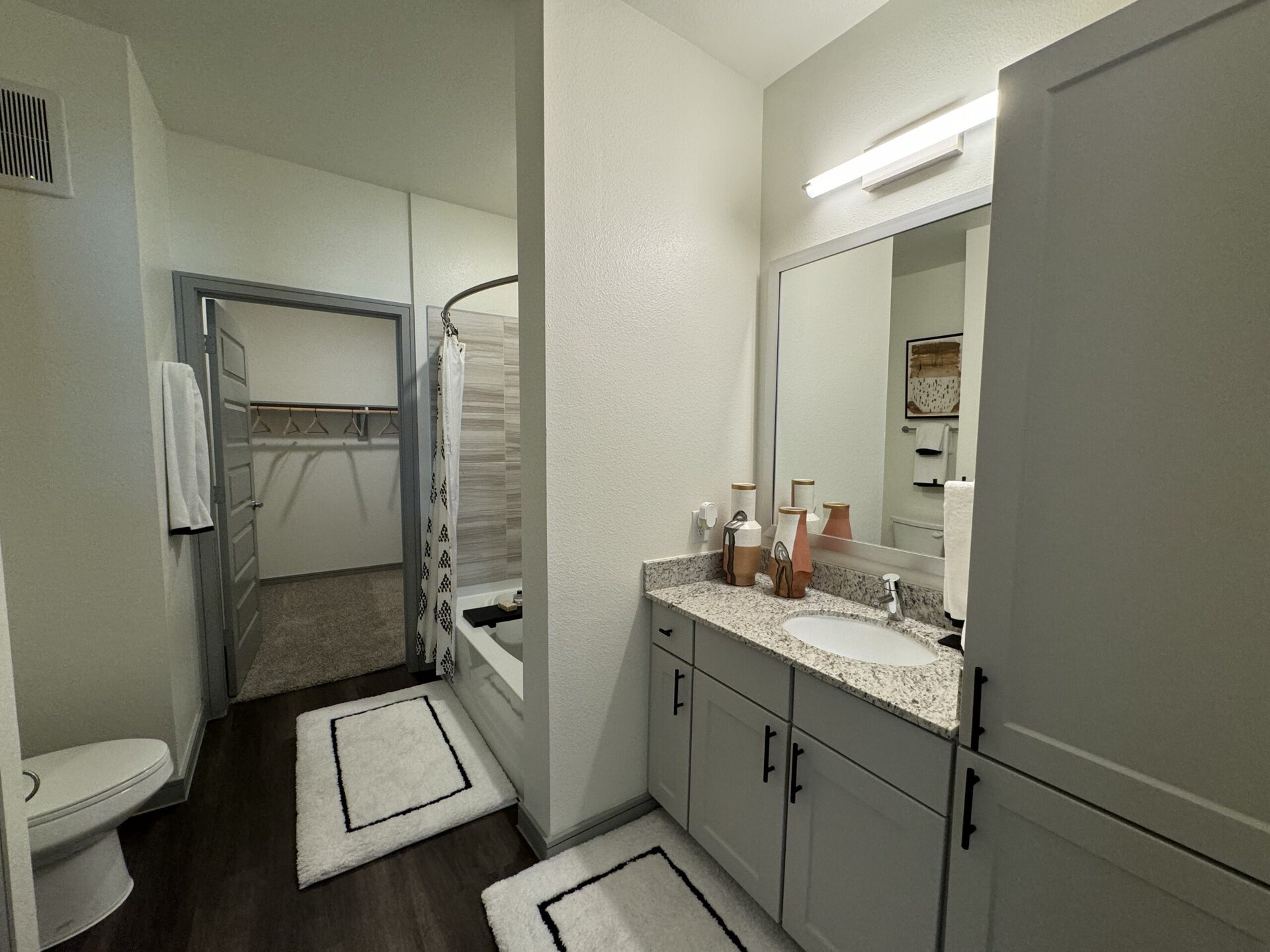 A modern bathroom features a light-colored granite countertop with an oval sink and stylish faucets. Above the sink, a large mirror reflects the space. Decorative vases in varying heights and colors are placed on the countertop. To the left, a shower area is enclosed with a patterned curtain, and a bathtub is visible. A towel rack with white towels hangs nearby. The floor is covered with a plush white rug, and a door leads to a closet with hanging space. The walls are painted in a soft, neutral tone, enhancing the bright and airy feel of the room.