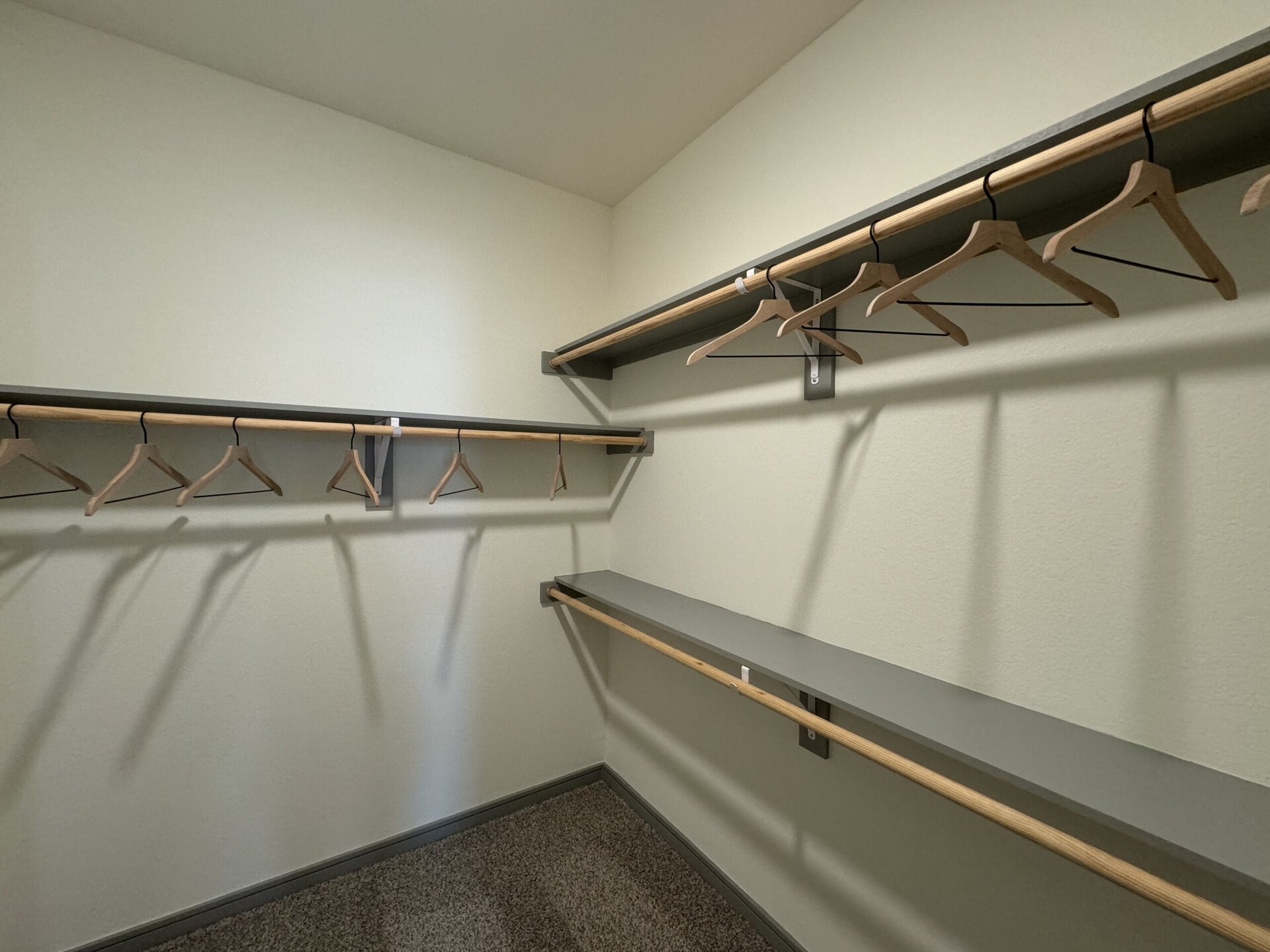 A spacious closet features two horizontal shelves and a hanging rod. The shelves are made of a smooth gray material with wooden accents, while the hanging rod is also wooden. Several wooden hangers are neatly arranged on the rod, casting shadows on the wall. The walls are painted a light color, contributing to a clean and organized appearance. The floor is carpeted in a neutral tone.