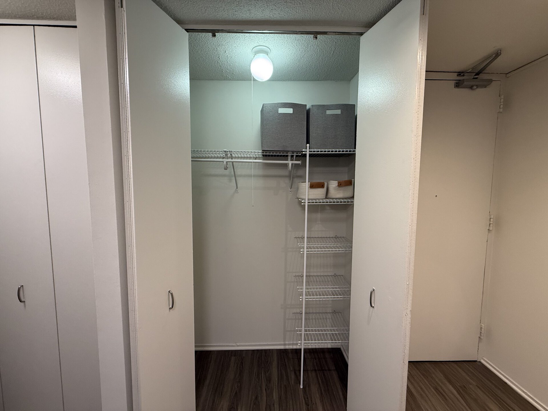 A small, organized closet features white walls and a light fixture hanging from the ceiling. On the left, there are two closed white doors. Inside the closet, a wire shelving unit is mounted on the back wall, holding two gray storage boxes and several white bowls with wooden handles. The shelving unit has multiple levels for additional storage, and the floor is covered with dark wood-like flooring.