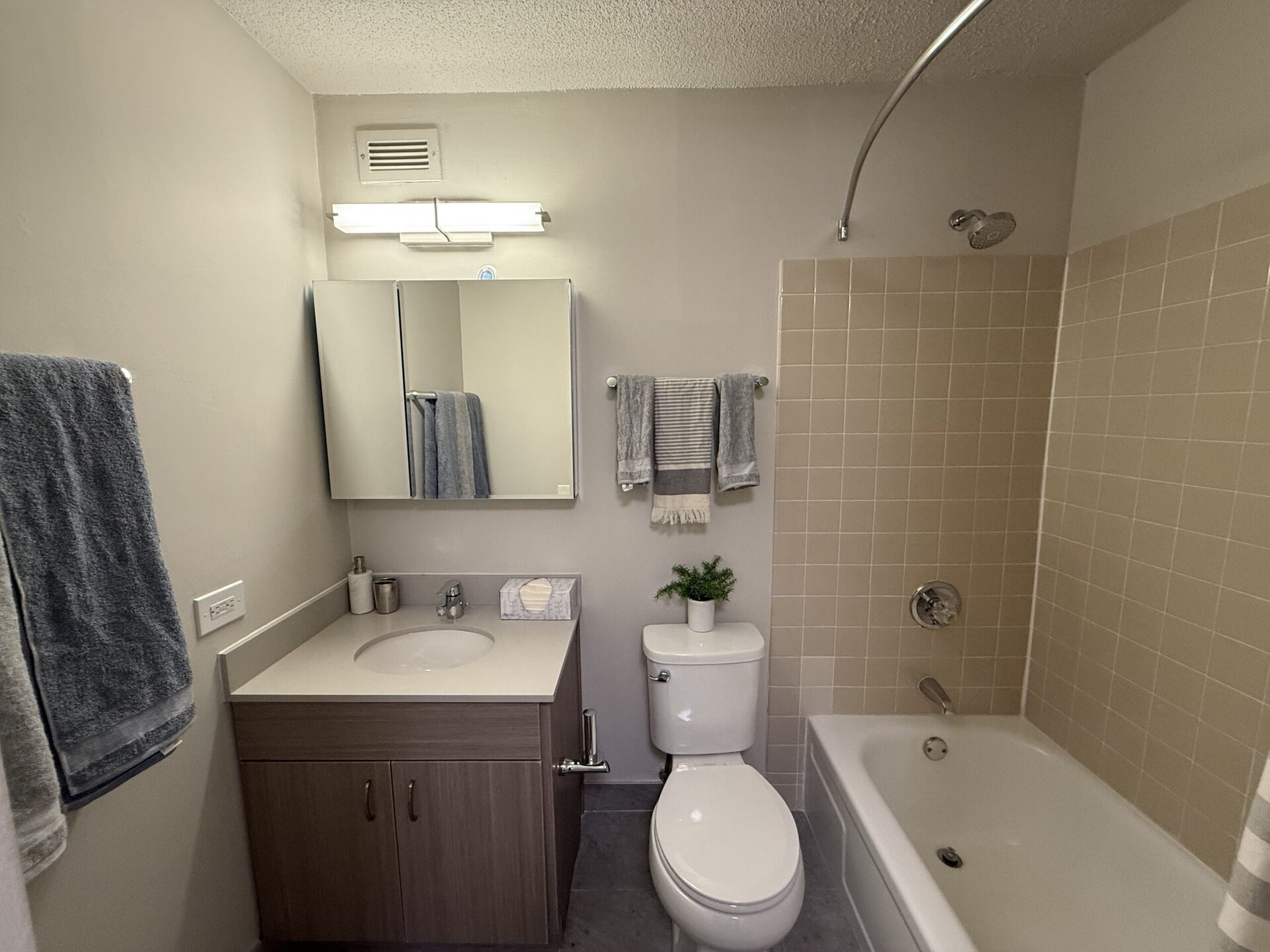 A modern bathroom featuring a light-colored tiled wall and a bathtub with a showerhead. To the left, there is a vanity with a round sink, a marble tissue box, and a small potted plant. Above the vanity, a three-panel mirror reflects the space. A towel rack holds two gray towels, and a striped hand towel hangs nearby. The toilet is positioned next to the vanity, completing the compact layout. The overall color scheme is neutral, creating a clean and inviting atmosphere.