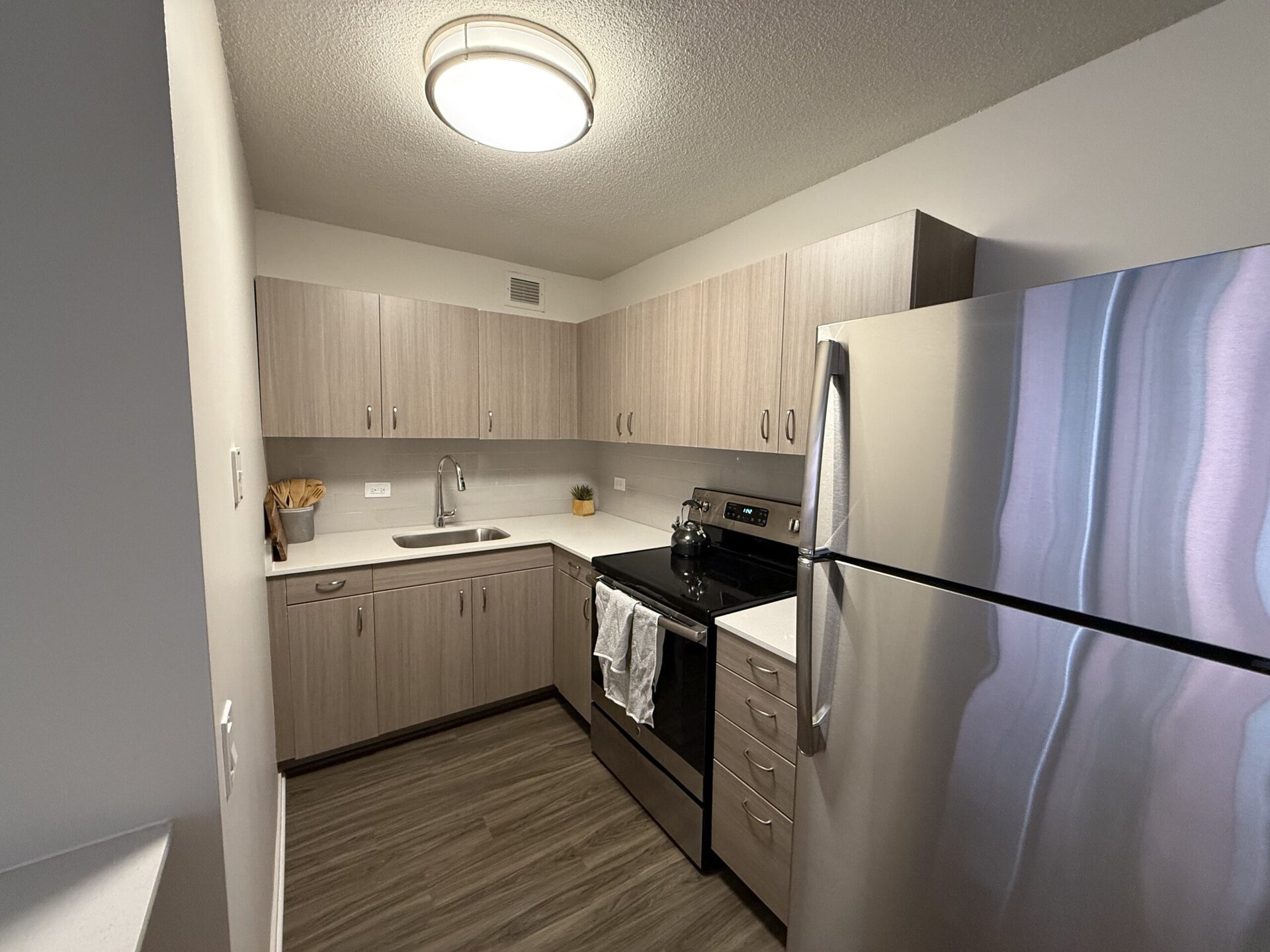 A modern kitchen featuring light wood cabinetry and a sleek design. The countertop is white with a stainless steel sink and a single-handle faucet. An electric stove with an oven is positioned next to the sink, and a stainless steel refrigerator stands on the right. A small potted plant adds a touch of greenery on the countertop, and a kitchen towel hangs from the oven handle. The flooring is a dark wood laminate, enhancing the contemporary feel of the space.