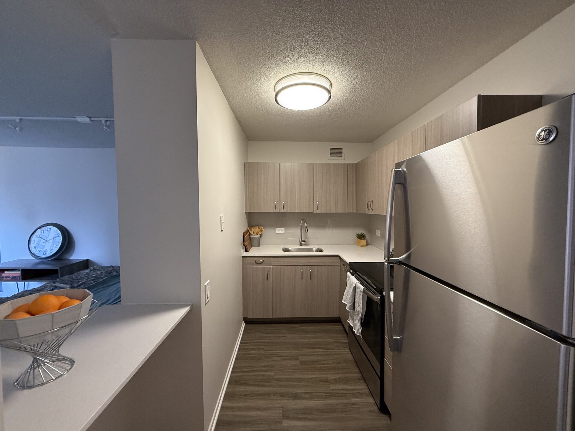 A modern kitchen features light wood cabinetry and a stainless steel refrigerator. The countertop is white with a small sink and a sleek faucet. A bowl of oranges sits on the counter, and there are kitchen utensils in a container nearby. In the background, a living area is visible with a large wall clock and a couch. The flooring is a dark wood laminate, and the space is well-lit by a ceiling fixture.