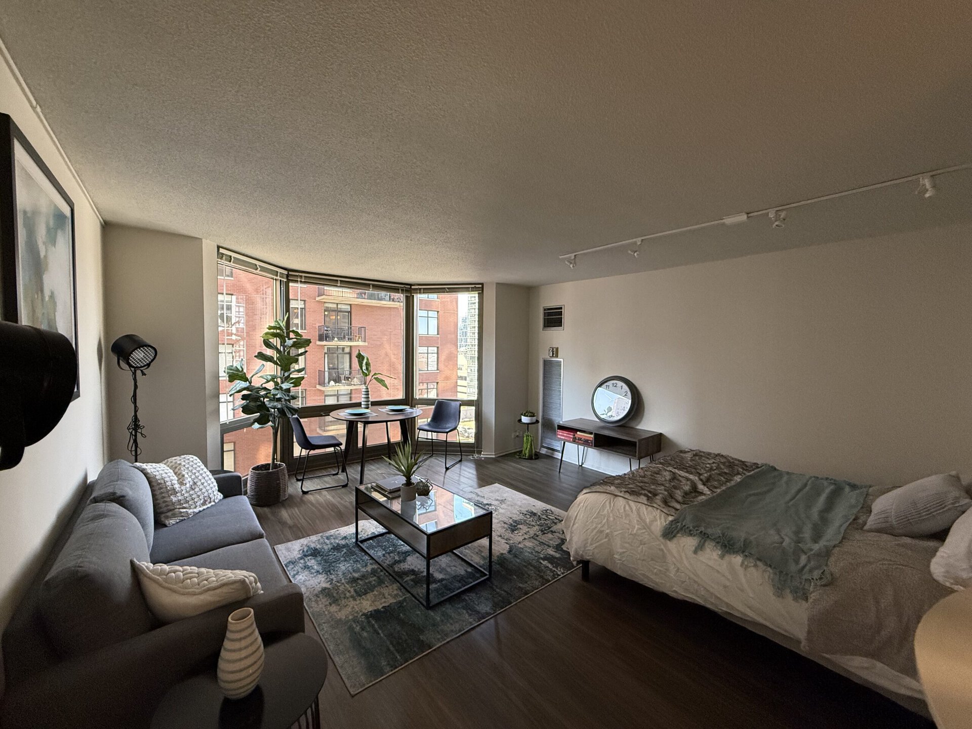 A modern studio apartment features a cozy gray sofa with decorative pillows, a glass coffee table on a patterned rug, and a bed with a light-colored blanket. A small dining area with two chairs and a table is positioned near large windows, allowing natural light to fill the space. A potted plant adds a touch of greenery, while a round wall clock and a small shelf complete the decor. The walls are painted in neutral tones, enhancing the airy feel of the room.