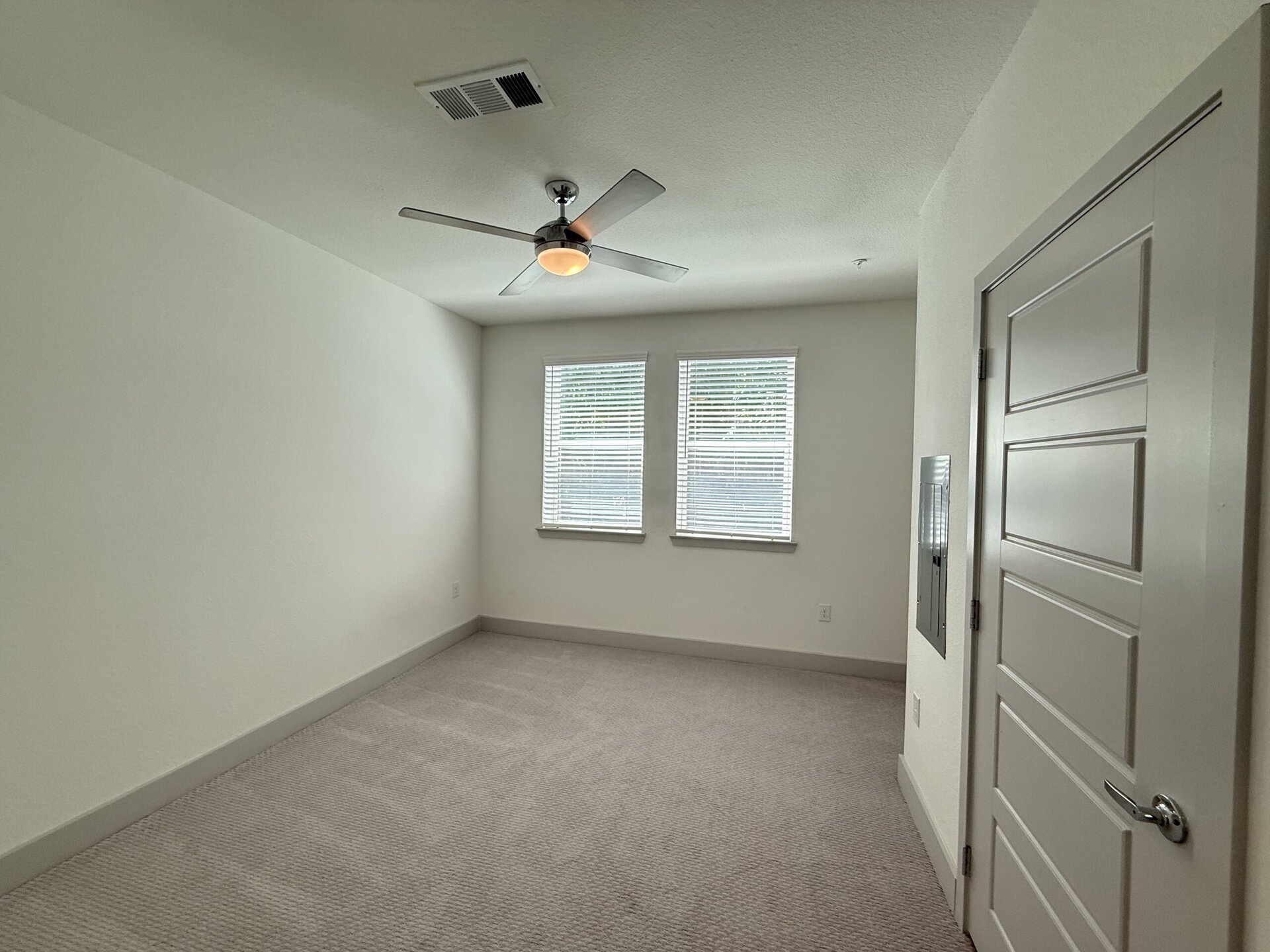A small, empty room features light-colored walls and a plush carpeted floor. Two windows with horizontal blinds allow natural light to enter, illuminating the space. A modern ceiling fan with a light fixture is mounted in the center of the ceiling. To the right, there is a closed door with a simple design, and next to it, a wall-mounted mailbox or intercom unit. The overall atmosphere is bright and minimalistic.