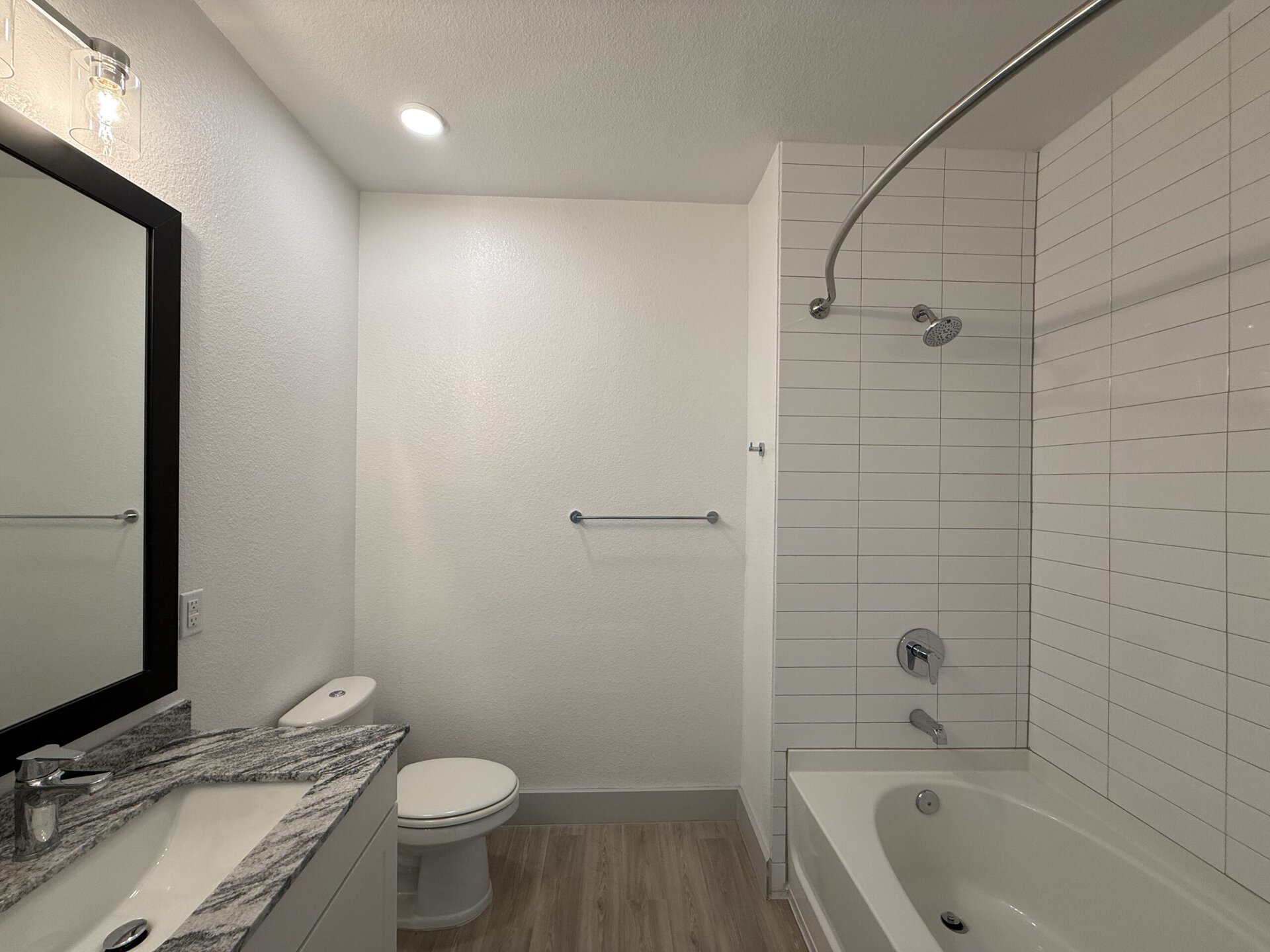 A modern bathroom features a white tiled shower area with a curved shower rod and a rainfall showerhead. To the left, there is a sleek vanity with a granite countertop, a rectangular sink, and a polished faucet. Above the sink, a large mirror with a black frame reflects the space. A toilet is positioned nearby, and a towel bar is mounted on the wall. The overall color scheme is light and neutral, creating a clean and spacious feel.
