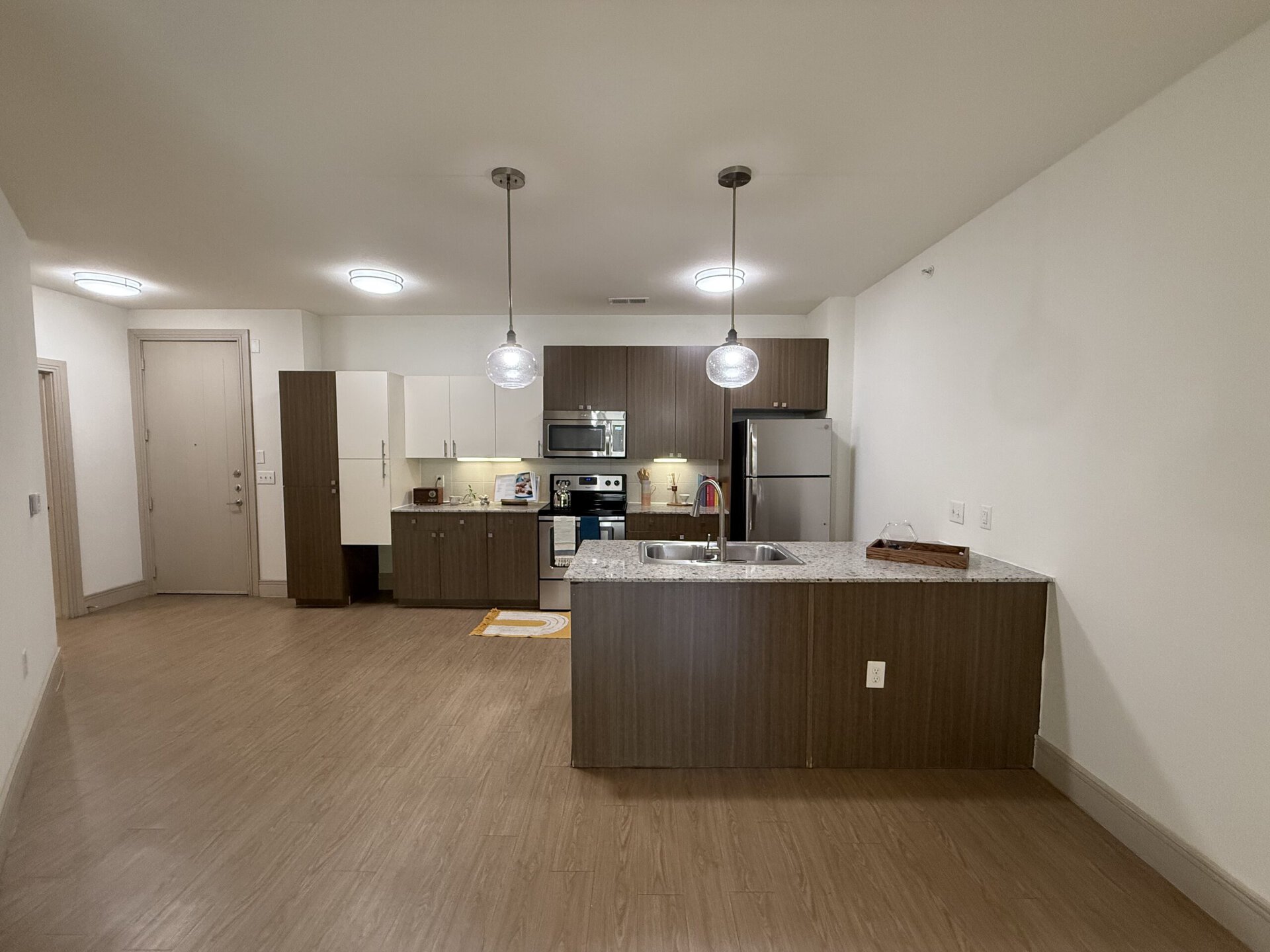 A modern kitchen space features a central island with a granite countertop and a double sink. Above the island, two pendant lights hang, providing illumination. The cabinetry is a mix of dark wood and white finishes, with a stainless steel refrigerator and oven visible. A small decorative tray sits on the countertop, and a rug with a circular design is placed on the floor. The walls are painted white, and the flooring is a light wood laminate. A door leads to the entrance of the apartment.