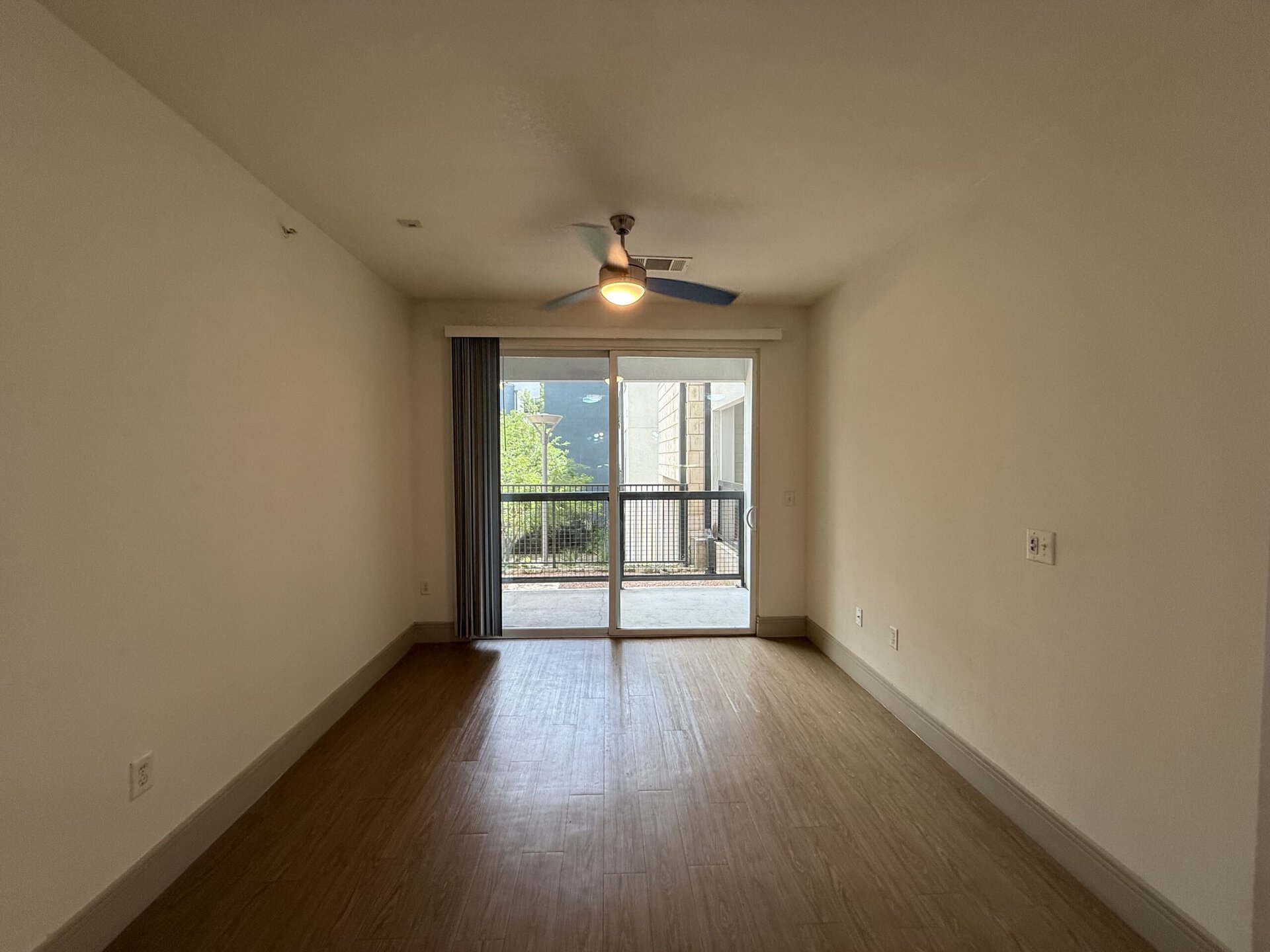 A spacious room with light-colored walls and a wooden floor. A ceiling fan with a light fixture is mounted in the center. Large sliding glass doors lead to a balcony, allowing natural light to fill the space. The balcony features a railing and greenery visible outside. The room is empty, providing a blank canvas for furnishings.