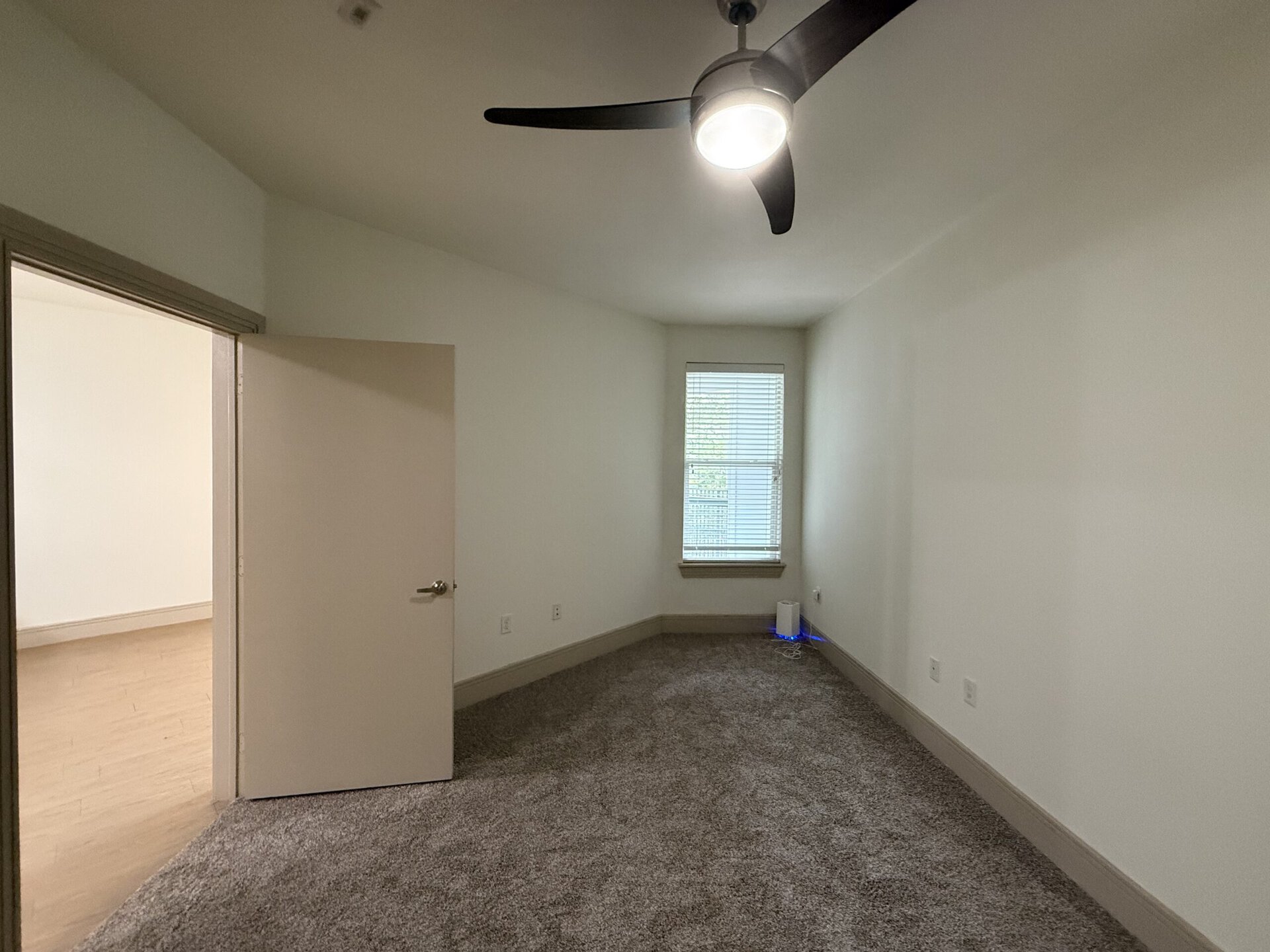 A small room features light-colored walls and a plush carpeted floor. A ceiling fan with a light fixture is installed above. To the left, there is an open door leading to another space. A window with blinds allows natural light to enter, and a small device with blue lighting is positioned on the floor near the wall. The overall atmosphere is bright and uncluttered.