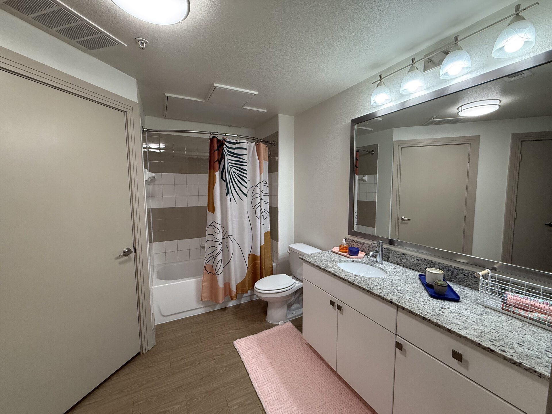 A modern bathroom features a bathtub with a shower, enclosed by a colorful shower curtain adorned with tropical leaves. The countertop is made of granite and includes a sink with a faucet, a small decorative tray with a cup and a plant, and a wire basket holding rolled towels. A large mirror above the sink reflects the space, while overhead lighting provides illumination. The floor is covered with a soft pink rug, and a door leads to another room.