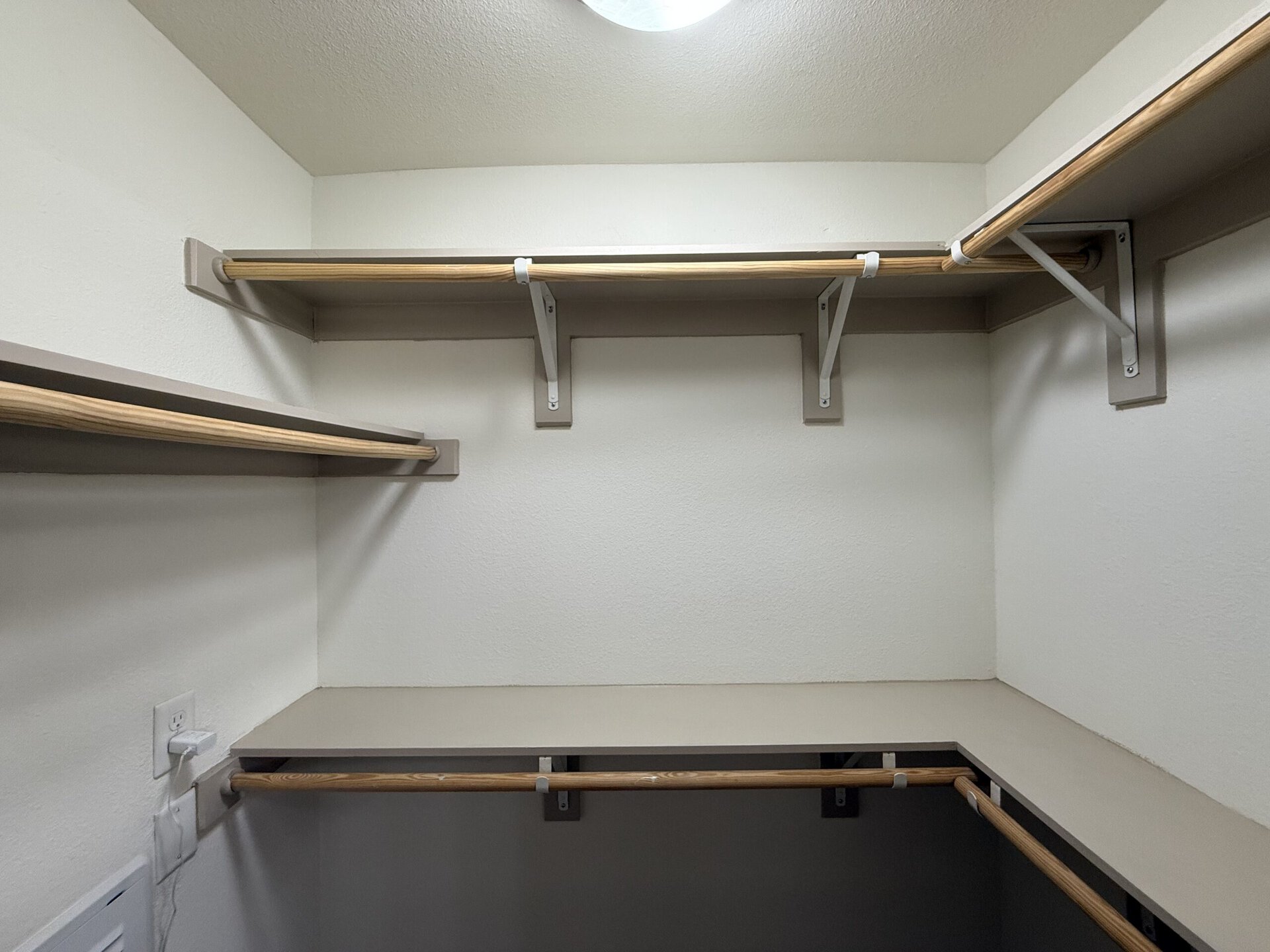 An empty closet features two wooden hanging rods supported by white brackets, positioned on the walls. Above the rods, there are two shelves, also supported by brackets, providing additional storage space. The walls are painted a light color, and there is a small countertop area at the bottom, with an electrical outlet visible on one side. The overall space is clean and organized, designed for efficient storage.