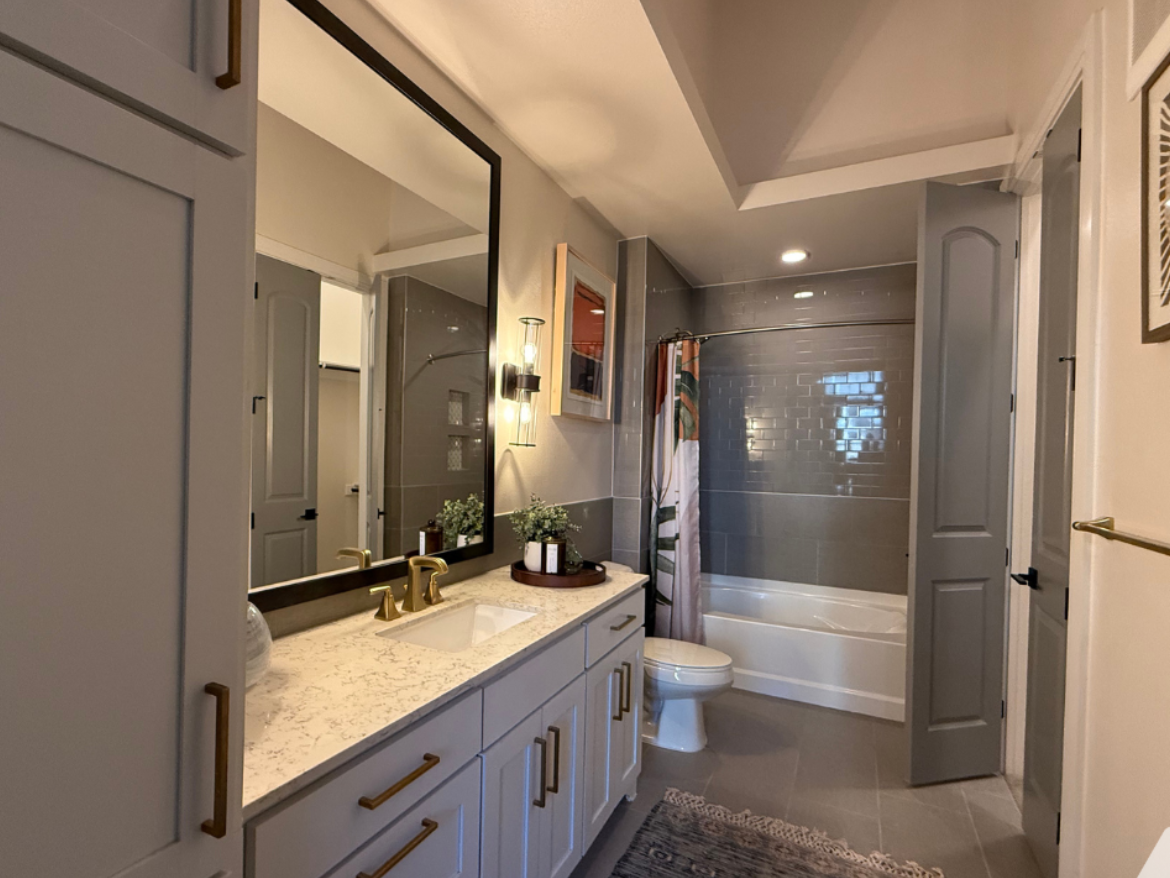 A modern bathroom features a light gray vanity with gold hardware and a white countertop. Above the sink, a large rectangular mirror is framed in black. To the right, a shower area is enclosed with gray tiles and a shower curtain displaying a leafy pattern. A white bathtub is positioned beneath the curtain. The space is well-lit with a wall-mounted light fixture and includes decorative elements like a small potted plant. The floor is tiled in a light gray, and a patterned rug adds warmth to the room.