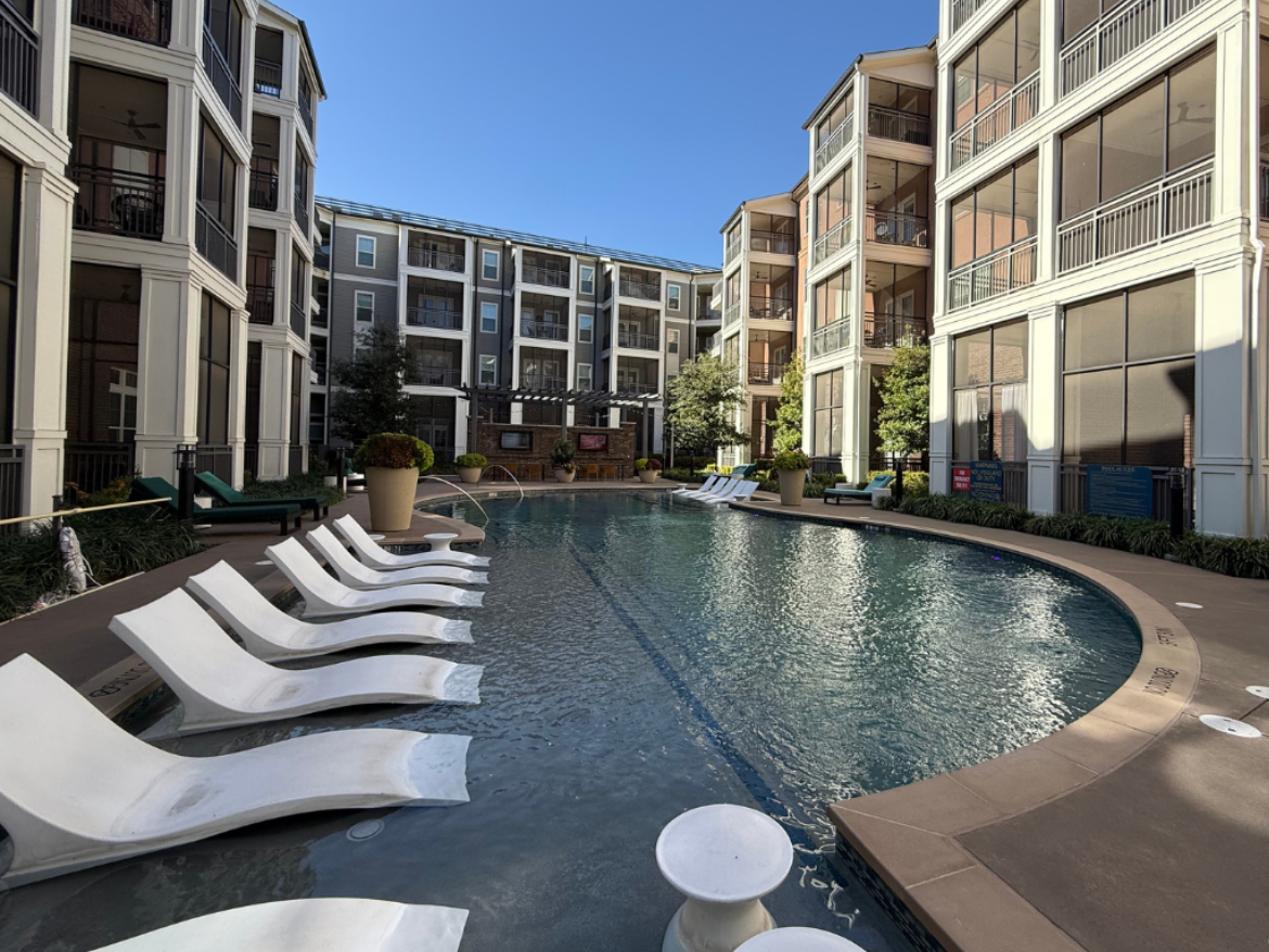 A modern swimming pool is situated in a courtyard surrounded by multi-story apartment buildings. The pool features a curved edge and is lined with several white lounge chairs positioned on a tan deck. Lush greenery and potted plants enhance the area, while the buildings have large windows and balconies. The sky is clear and blue, creating a bright and inviting atmosphere.