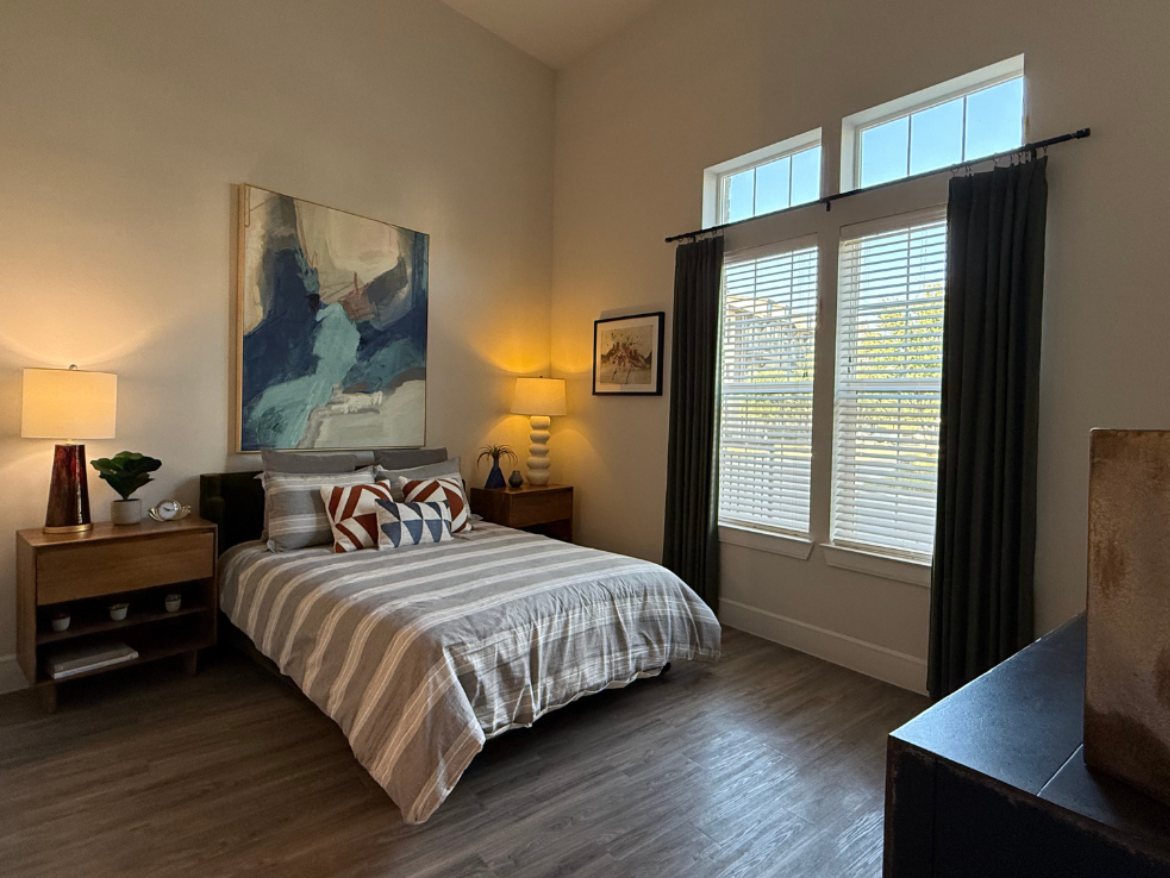 A cozy bedroom features a queen-sized bed with a striped gray and white duvet. Decorative pillows in various patterns, including geometric designs, are arranged on the bed. On either side of the bed are wooden nightstands, each with a lamp and a small plant. Above the bed hangs a large abstract painting with shades of blue and green. Two tall windows with blinds allow natural light to enter, framed by dark green curtains. A small framed artwork is mounted on the wall near the window, and a textured decorative object is visible on a nearby surface. The flooring is a warm wood tone, enhancing the room's inviting atmosphere.