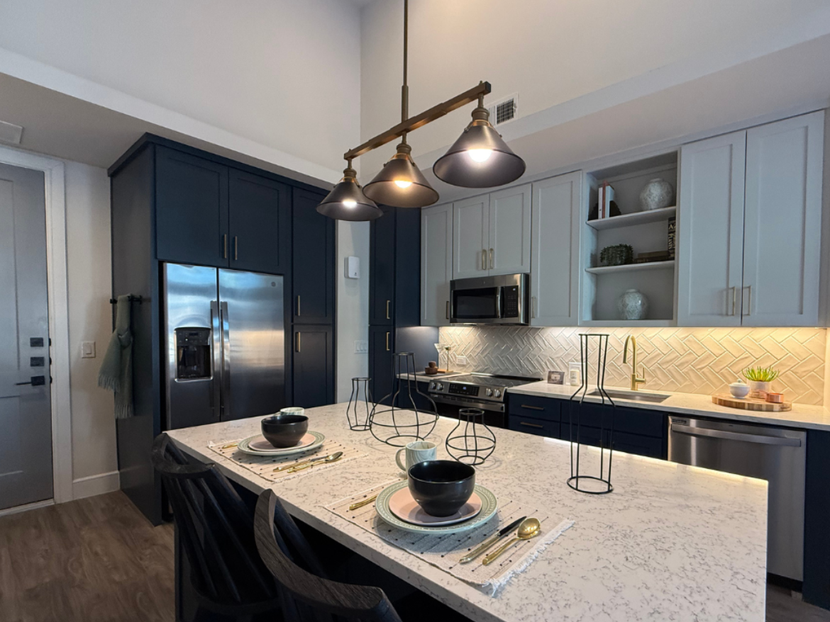 A modern kitchen features a large island with a white speckled countertop. On the island, there are two black bowls, plates, and gold utensils arranged neatly. Above the island, three pendant lights hang, providing illumination. The cabinetry is a mix of dark blue and light gray, with a herringbone-patterned backsplash behind the stove. Stainless steel appliances, including a refrigerator and microwave, are integrated into the design. A few decorative items are placed on open shelves, adding a touch of style. The flooring is a warm wood tone, enhancing the contemporary feel of the space.