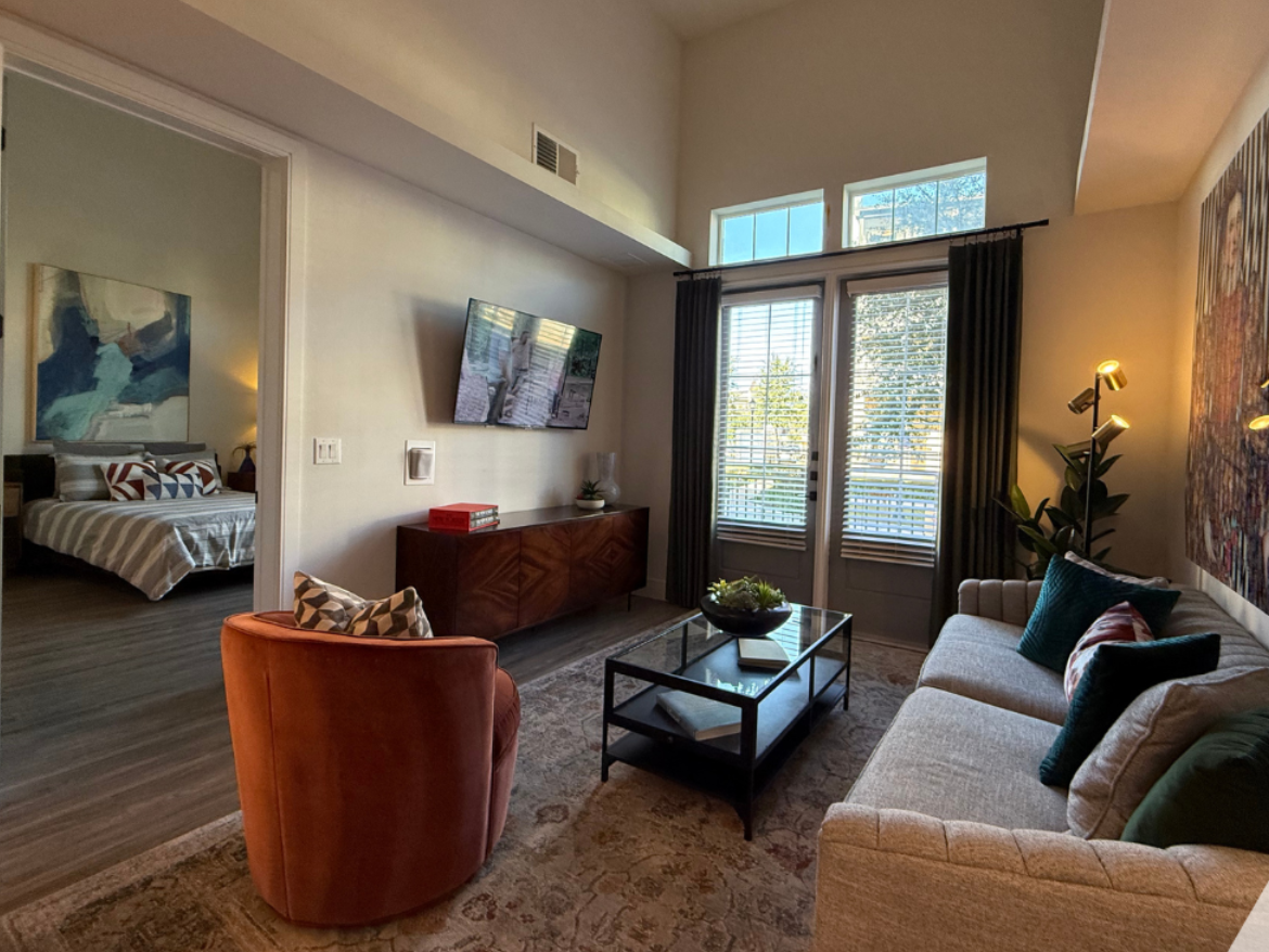A cozy living room features a light-colored sofa adorned with green and patterned pillows, paired with a round, orange accent chair. A glass coffee table sits in the center, displaying a decorative bowl and a few books. The wall opposite the sofa has a large flat-screen TV mounted above a wooden console. Natural light streams in through large windows with dark curtains, revealing a view of greenery outside. In the background, a bedroom is visible, showcasing a striped bedspread and abstract artwork on the wall. The flooring is a warm wood tone, enhancing the inviting atmosphere.