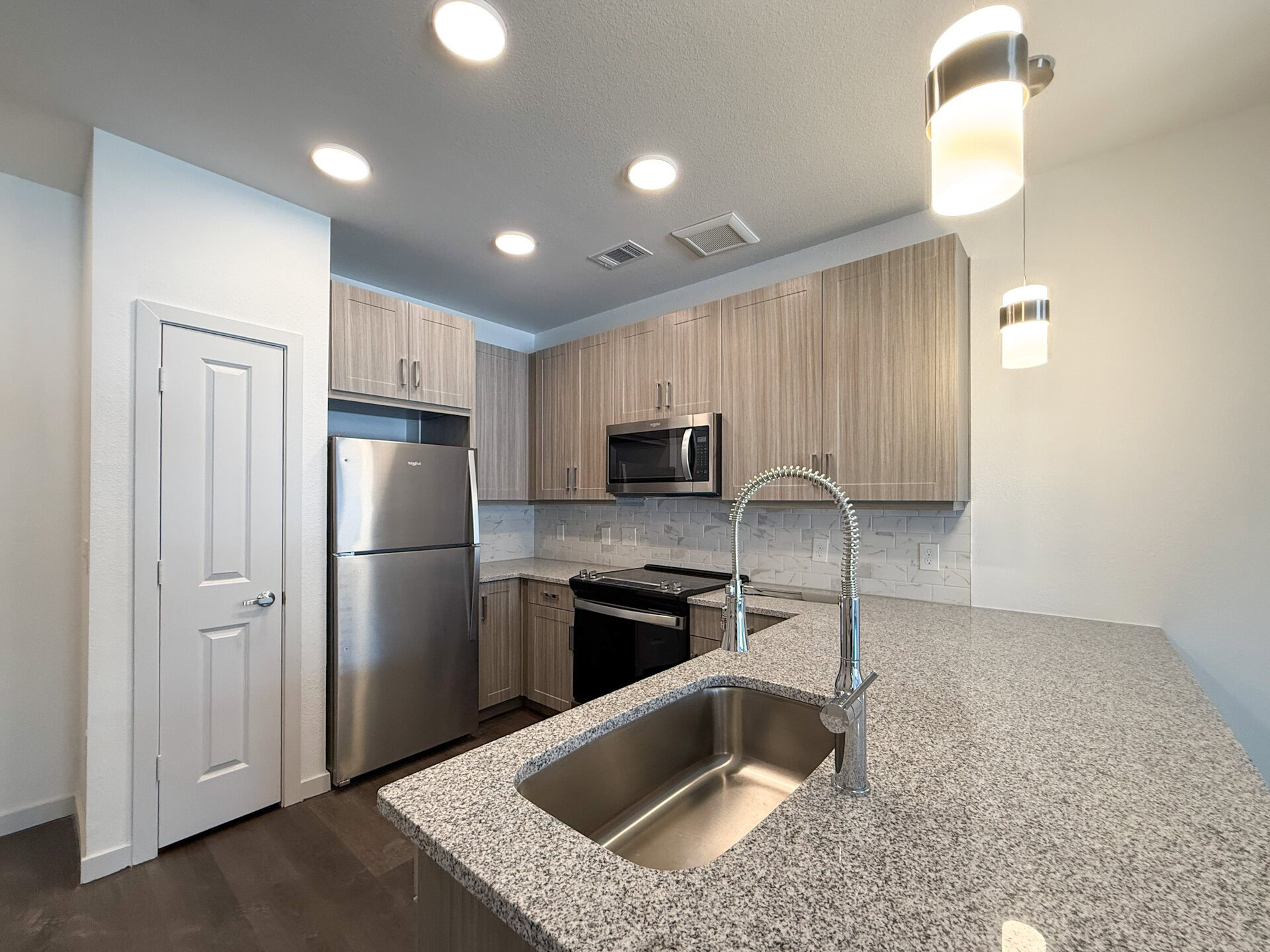 A modern San Antonio apartment kitchen featuring light wood cabinetry and a granite countertop. The stainless steel appliances include a refrigerator, microwave, and stove. A sleek, pull-down faucet is positioned over a stainless steel sink. The backsplash consists of white tiles, and the space is illuminated by recessed lighting and a pendant light.