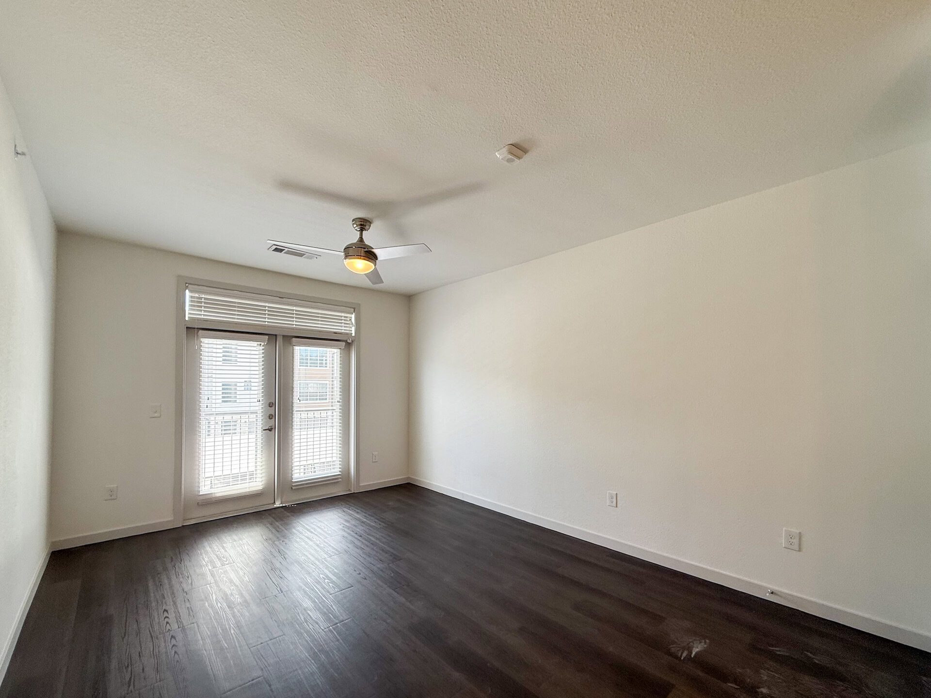A bright, San Antonio apartment living room features a ceiling fan with a light fixture. Two large balcony doors with blinds, allow natural light to fill the space. The overall atmosphere is clean and modern luxe.