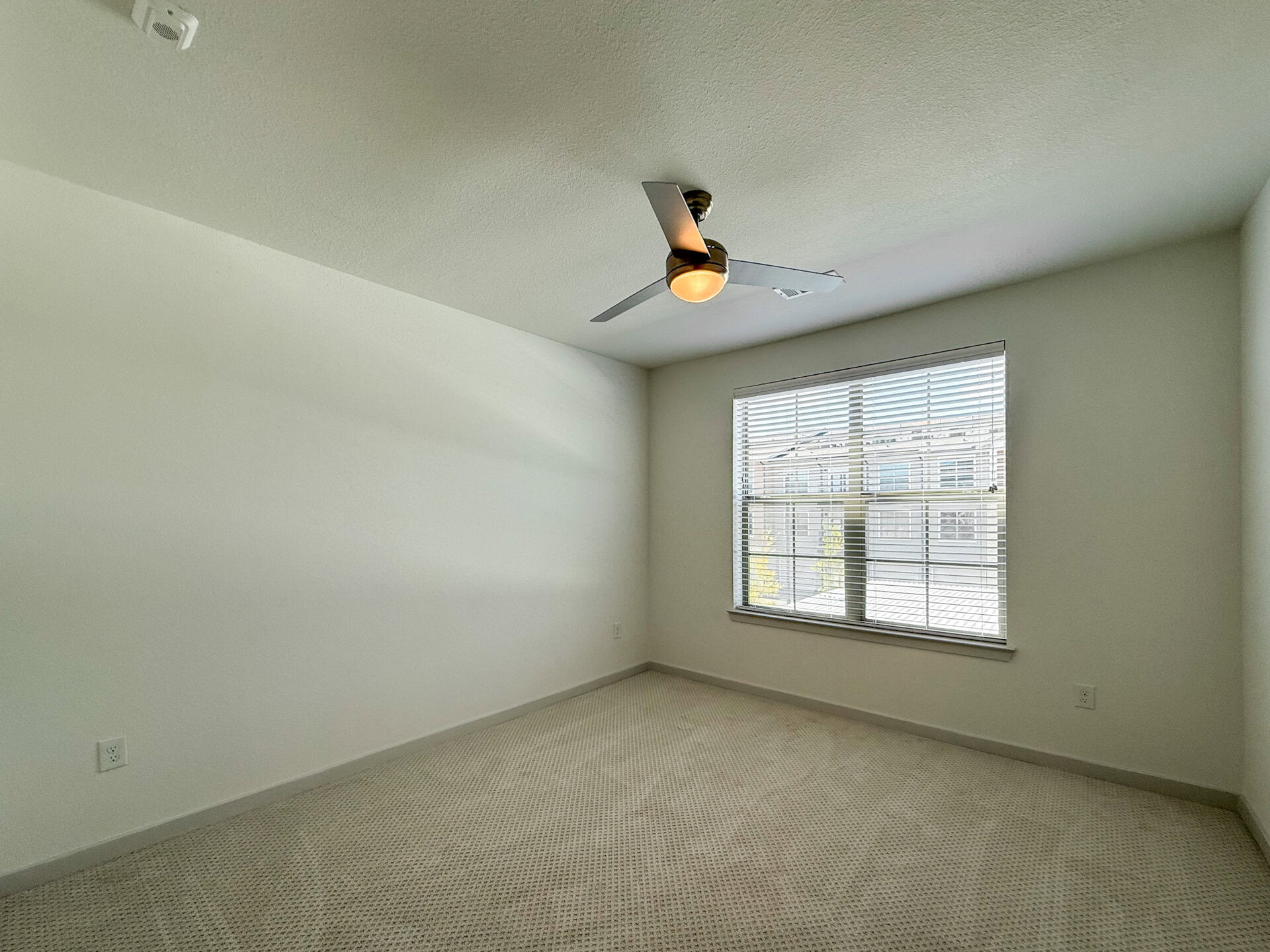 A bright, San Antonio apartment bedroom features light-colored walls and a ceiling fan with a light fixture. The room has a window with horizontal blinds, allowing natural light to enter. The floor is covered with a light carpet. The overall atmosphere is modern, clean, and minimalistic.