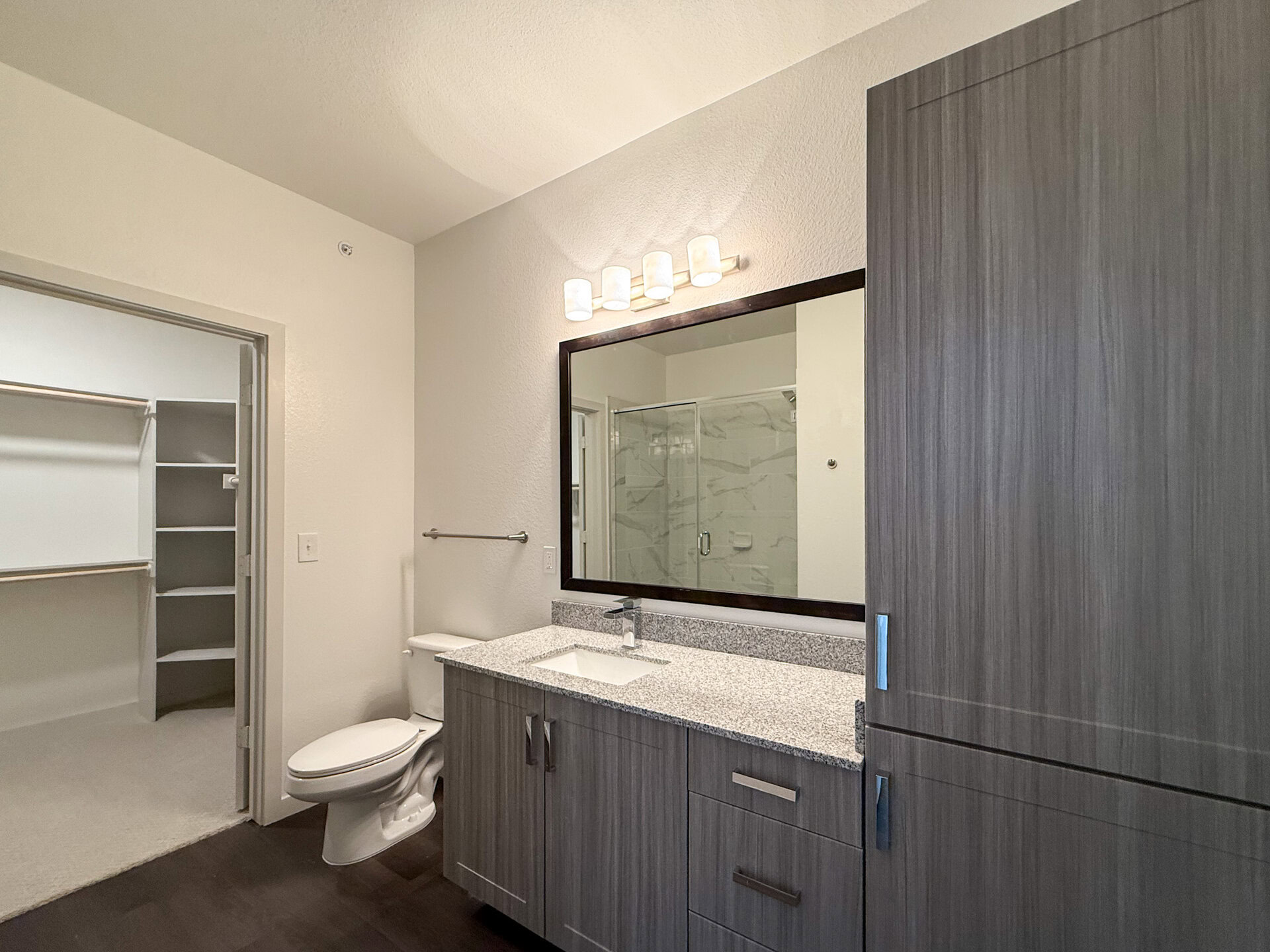 A modern San Antonio apartment bathroom features a sleek vanity with sand cabinets and light grey granite countertop and a rectangular sink. Above the vanity, a large mirror is framed in dark wood, complemented by four cylindrical light fixtures. To the left, a toilet is positioned next to a walk-in closet with built-in shelving. A glass shower enclosure is visible in the background, showcasing a marble-patterned tile design. The walls are painted in a light color, enhancing the spacious feel of the room.