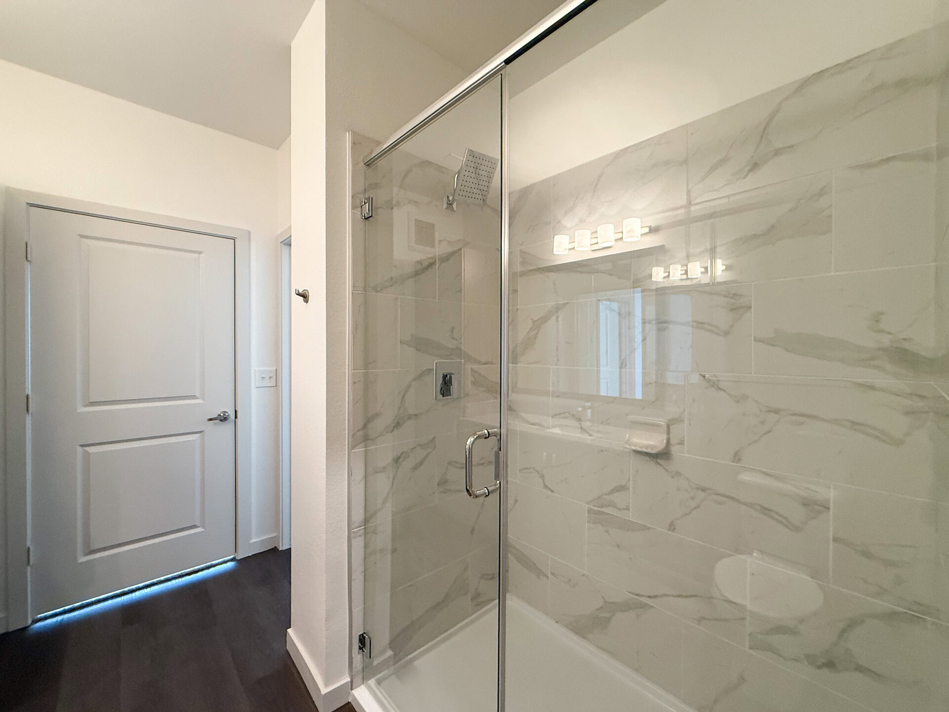 A modern San Antonio apartment spacious standing shower features a glass shower enclosure with a sleek chrome handle. The walls are adorned with large, light-colored tiles that have a marble-like pattern. A showerhead is mounted on the wall inside the shower. The overall aesthetic is clean and modern luxe.
