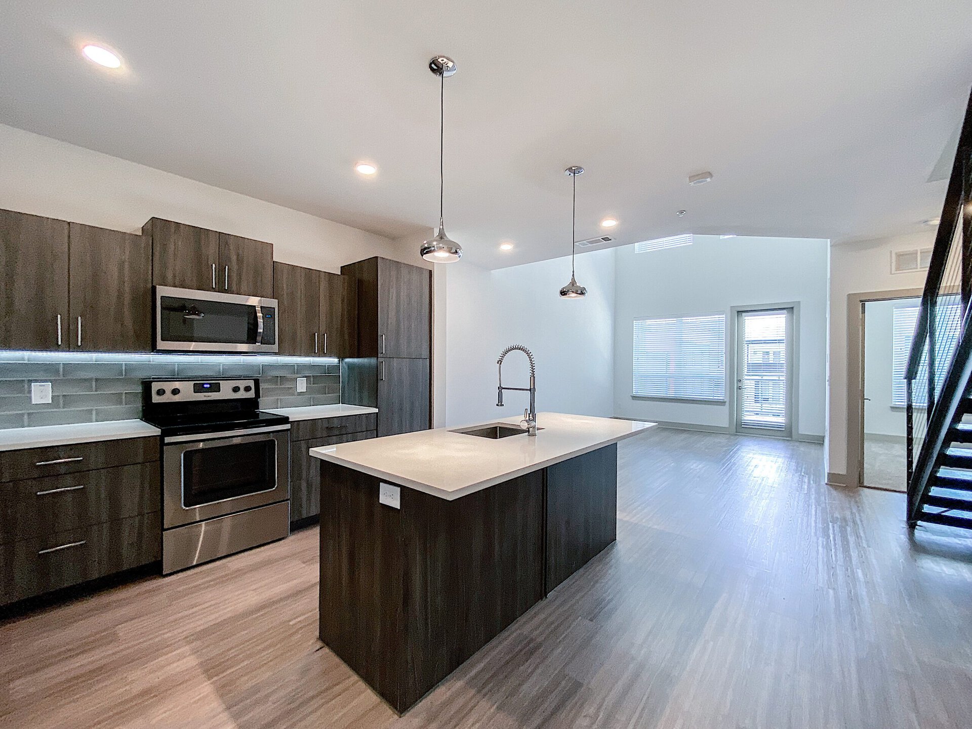 A modern luxe San Antonio loft apartment kitchen features dark grey wood cabinetry and stainless steel appliances, including an oven, microwave, and a sleek sink with a pull-down faucet. The countertop is light-colored and extends into a spacious island. The open layout leads to a living area with large windows allowing natural light, and a staircase is visible on the right side. The flooring is a light wood finish, enhancing the contemporary feel of the space.