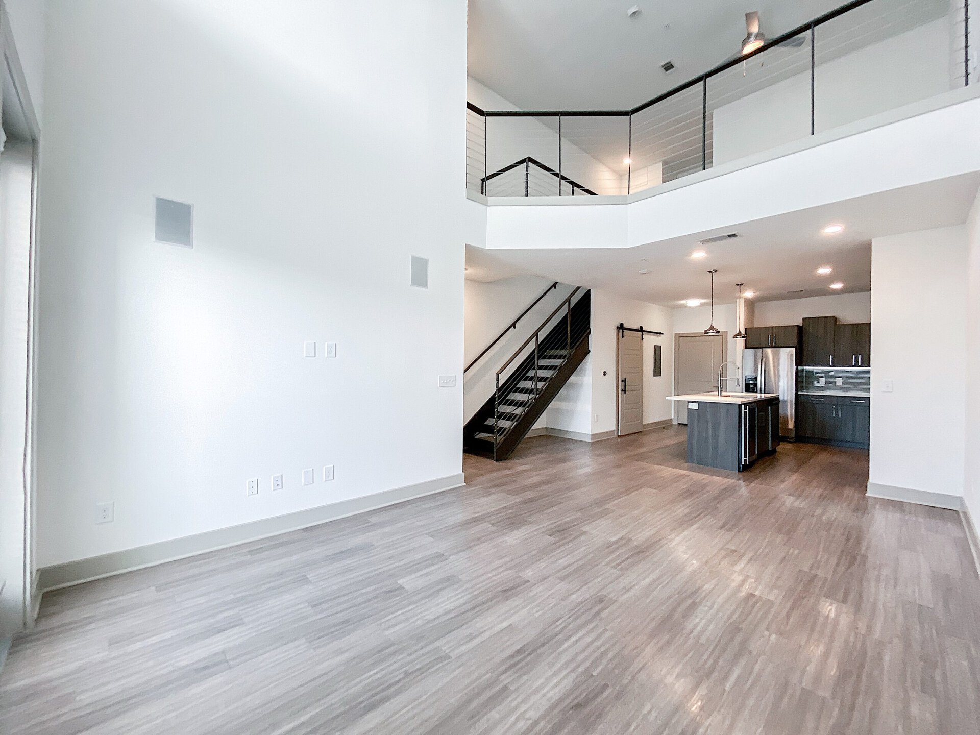 A spacious, modern luxe San Antonio loft apartment living area features light-colored walls and a wood-like floor. To the right, a contemporary kitchen with dark grey cabinetry and stainless steel appliances is visible, including an island with a sink. A staircase with a sleek railing leads to a lofted area above. The room is well-lit with recessed lighting and large windows, creating an open and airy atmosphere.