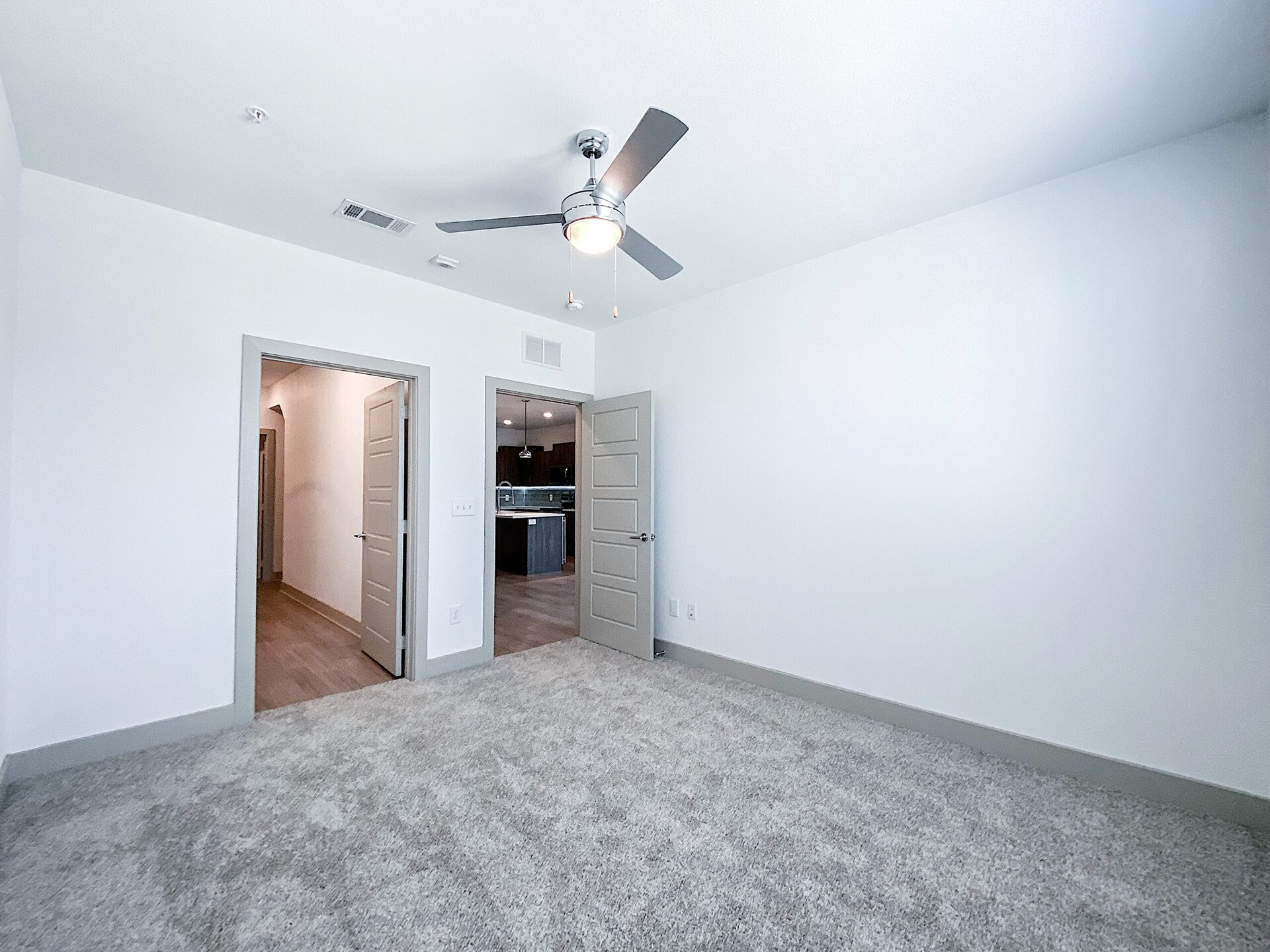 A spacious San Antonio loft apartment bedroom with light gray walls and plush carpet flooring. A ceiling fan with a light fixture is mounted above. The overall ambiance is bright and inviting.