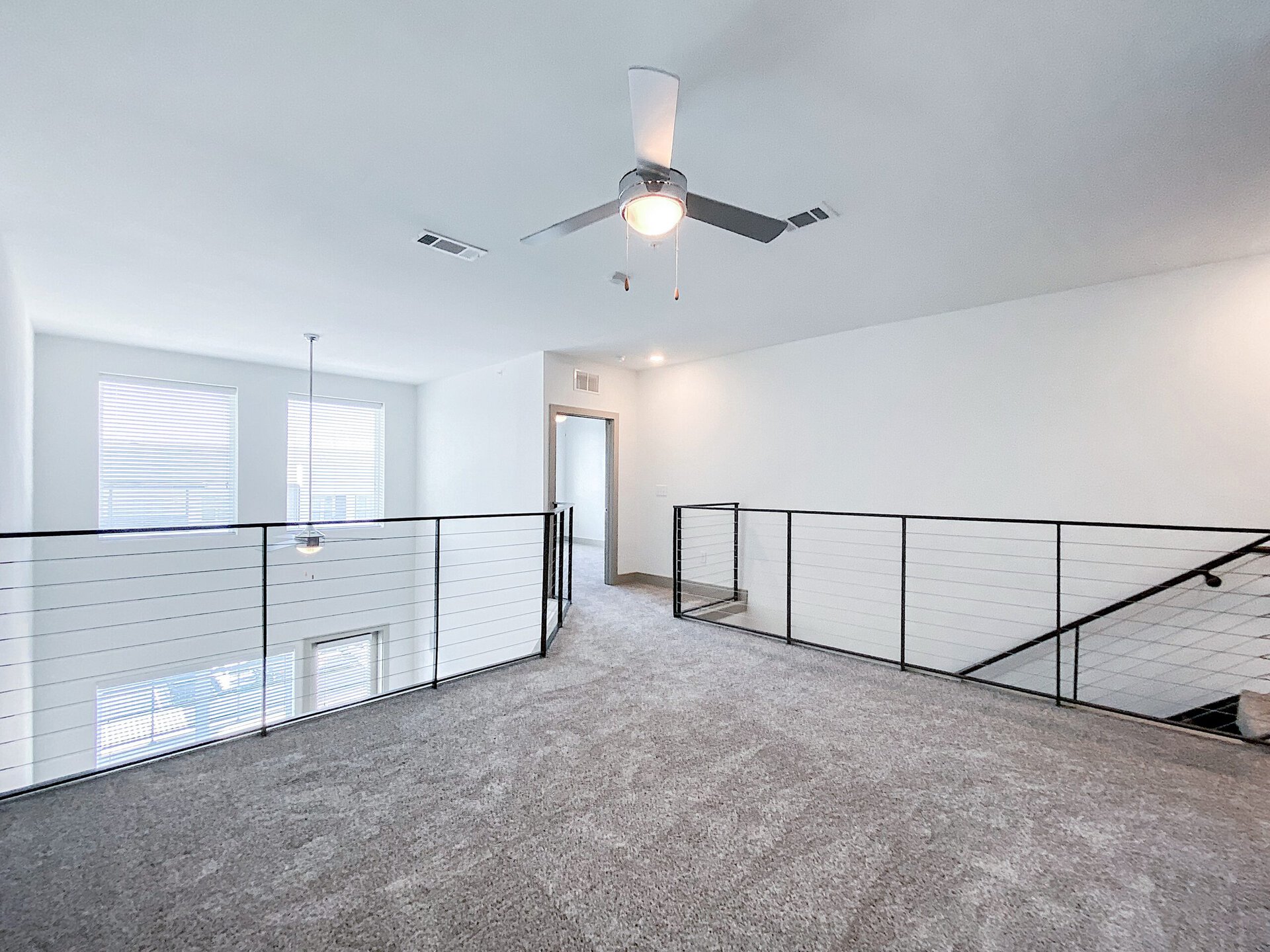 A spacious, well-lit San Antonio two bed apartment loft area features a light gray carpet and white walls. Two large windows with blinds allow natural light to fill the space. A modern ceiling fan hangs from the ceiling, and a pendant light is suspended above the open area. The loft is bordered by sleek black railings, leading to a staircase on one side.