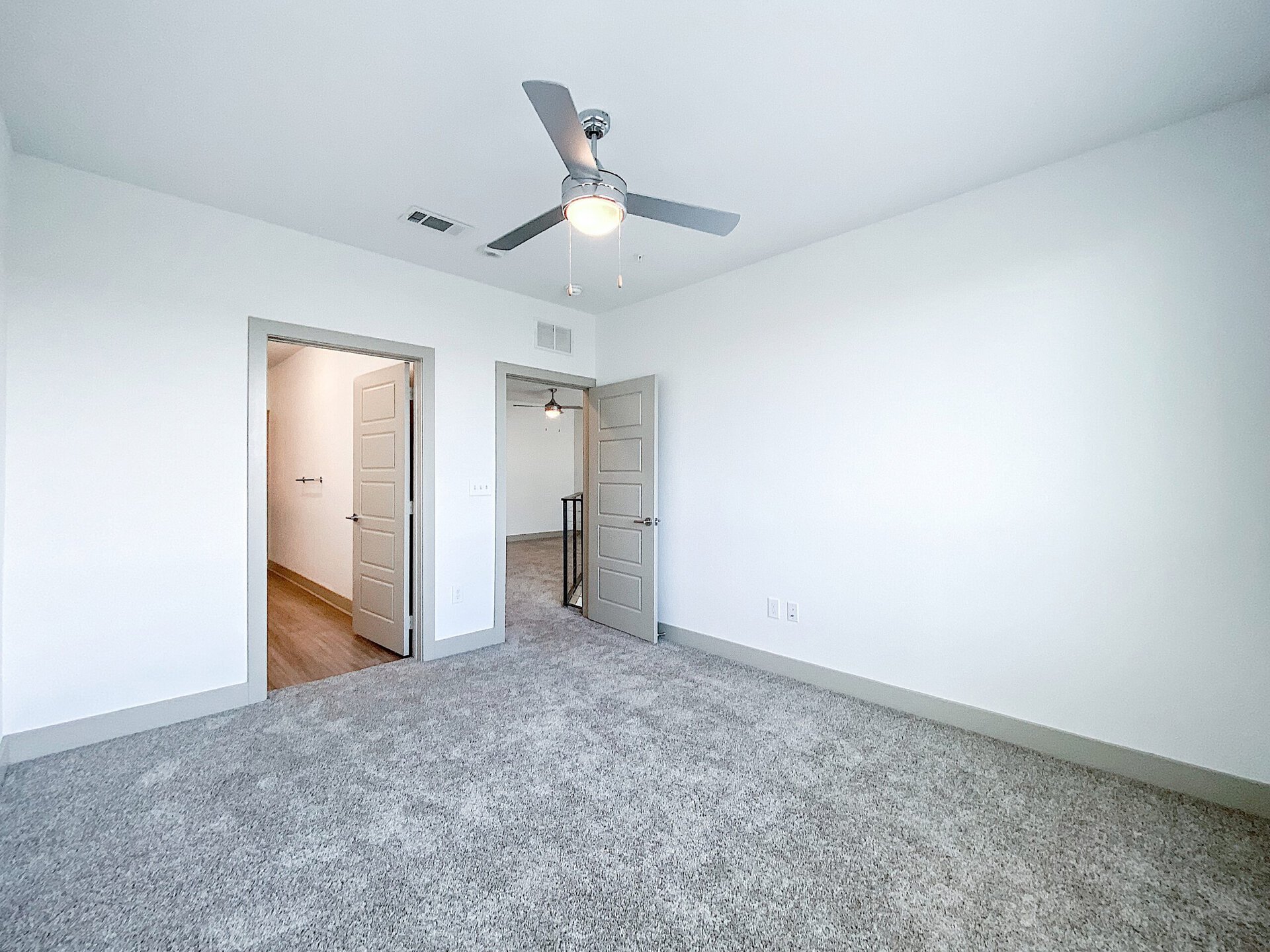 A spacious San Antonio loft apartment bedroom with light gray walls and plush carpet flooring. A ceiling fan with a light fixture is mounted above. The overall ambiance is bright and inviting.