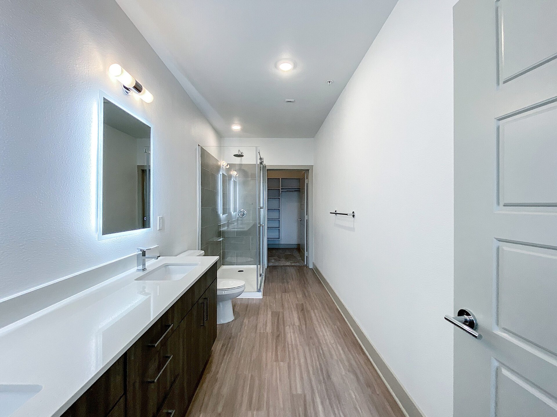 A modern luxe San Antonio loft apartment bathroom features a sleek, white countertop with a rectangular sink and a chrome faucet. Above the sink, a backlit mirror is mounted on the wall. To the left, a glass shower enclosure showcases tiled walls and a showerhead. The floor is covered in light wood-like laminate, and a toilet is positioned beside the sink. A towel bar is mounted on the wall, and a door leads to a closet space in the background. The walls are painted white, creating a bright and airy atmosphere.