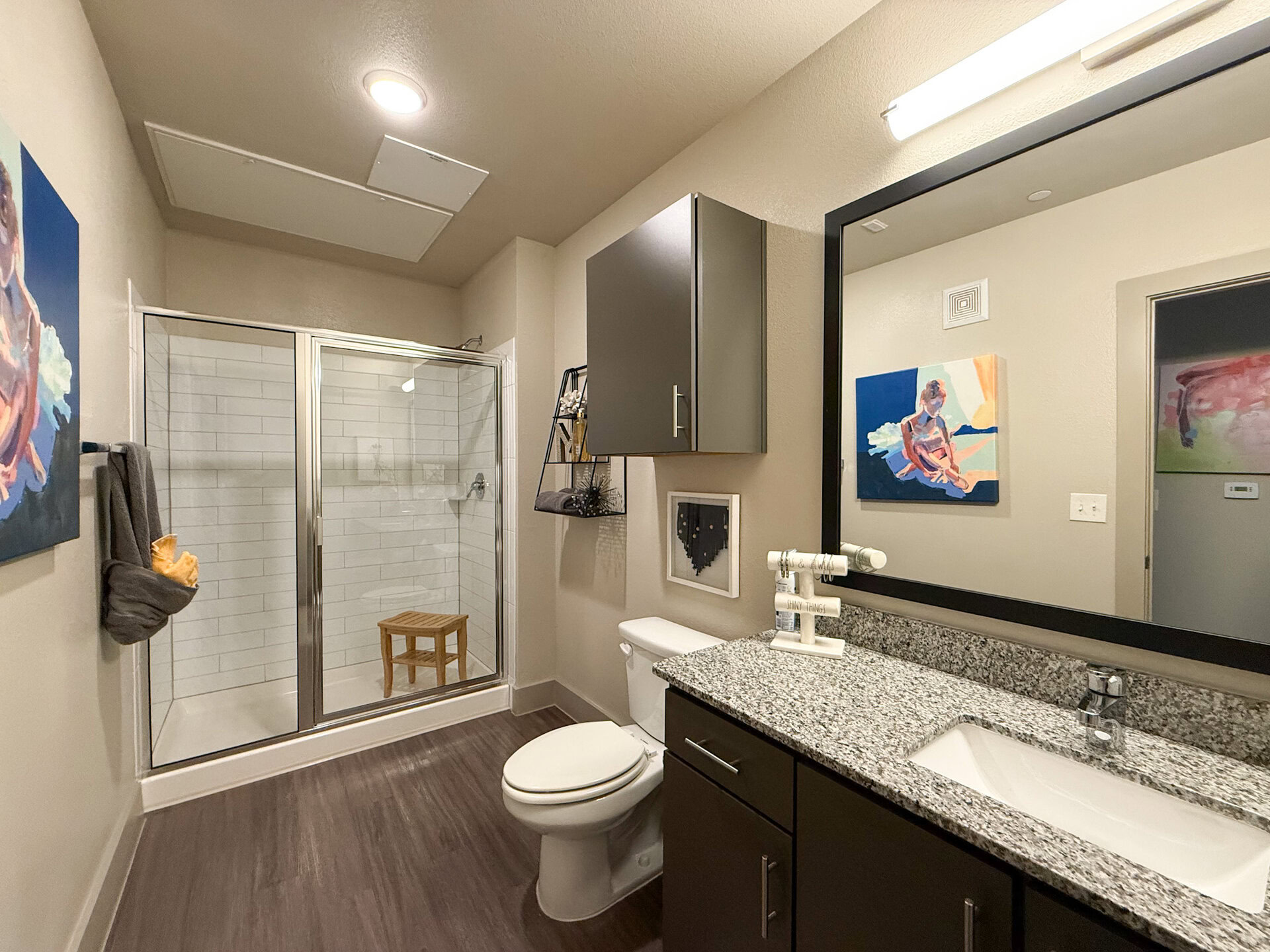A modern San Antonio apartment bathroom featuring a glass shower enclosure with white tiled walls. The space includes a dark cabinet above a granite countertop with a sink and faucet. The flooring is dark wood, contributing to a modern aesthetic.