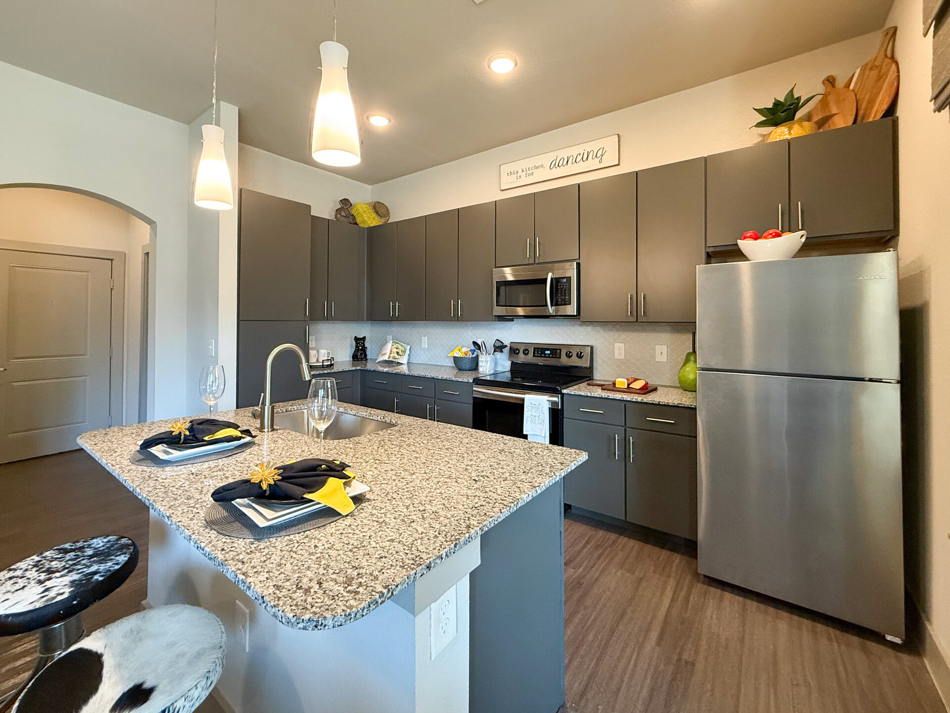 A modern San Antonio apartment kitchen featuring dark cabinetry and a spacious granite countertop island. Stainless steel appliances, including a refrigerator and oven. The flooring is a warm wood tone, enhancing the inviting atmosphere.