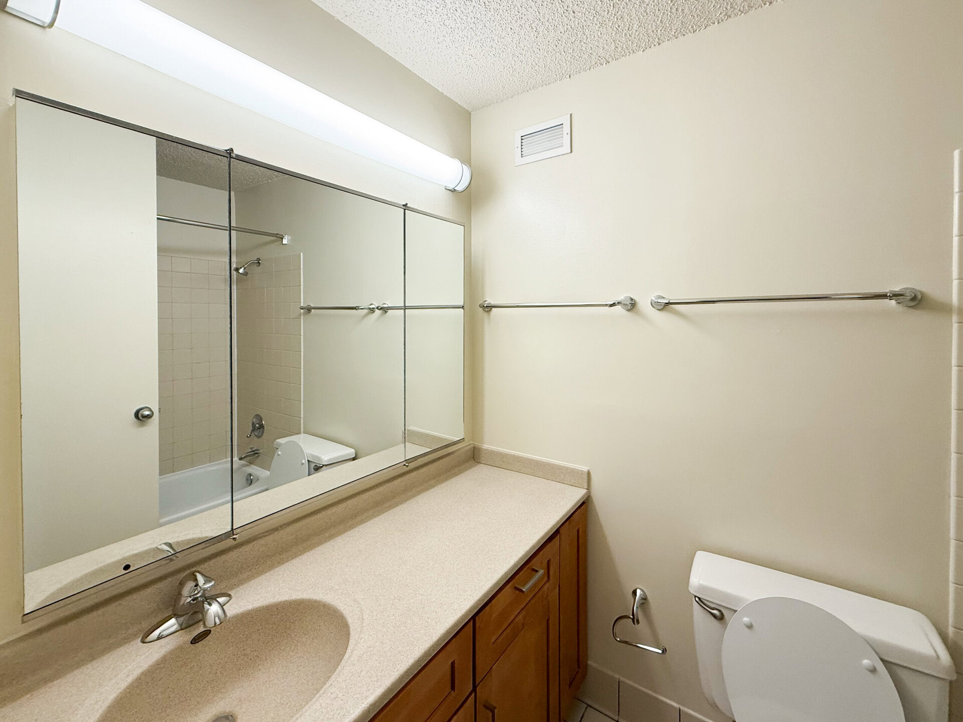 A bathroom features a long, mirrored wall above a beige countertop with a sink. The countertop is smooth and has a faucet. To the right, a white toilet is visible, and a shower area with tiled walls is partially seen in the background. Two towel bars are mounted on the wall, and a light fixture runs along the top of the mirror. The walls are painted a light color, contributing to a bright and clean appearance.