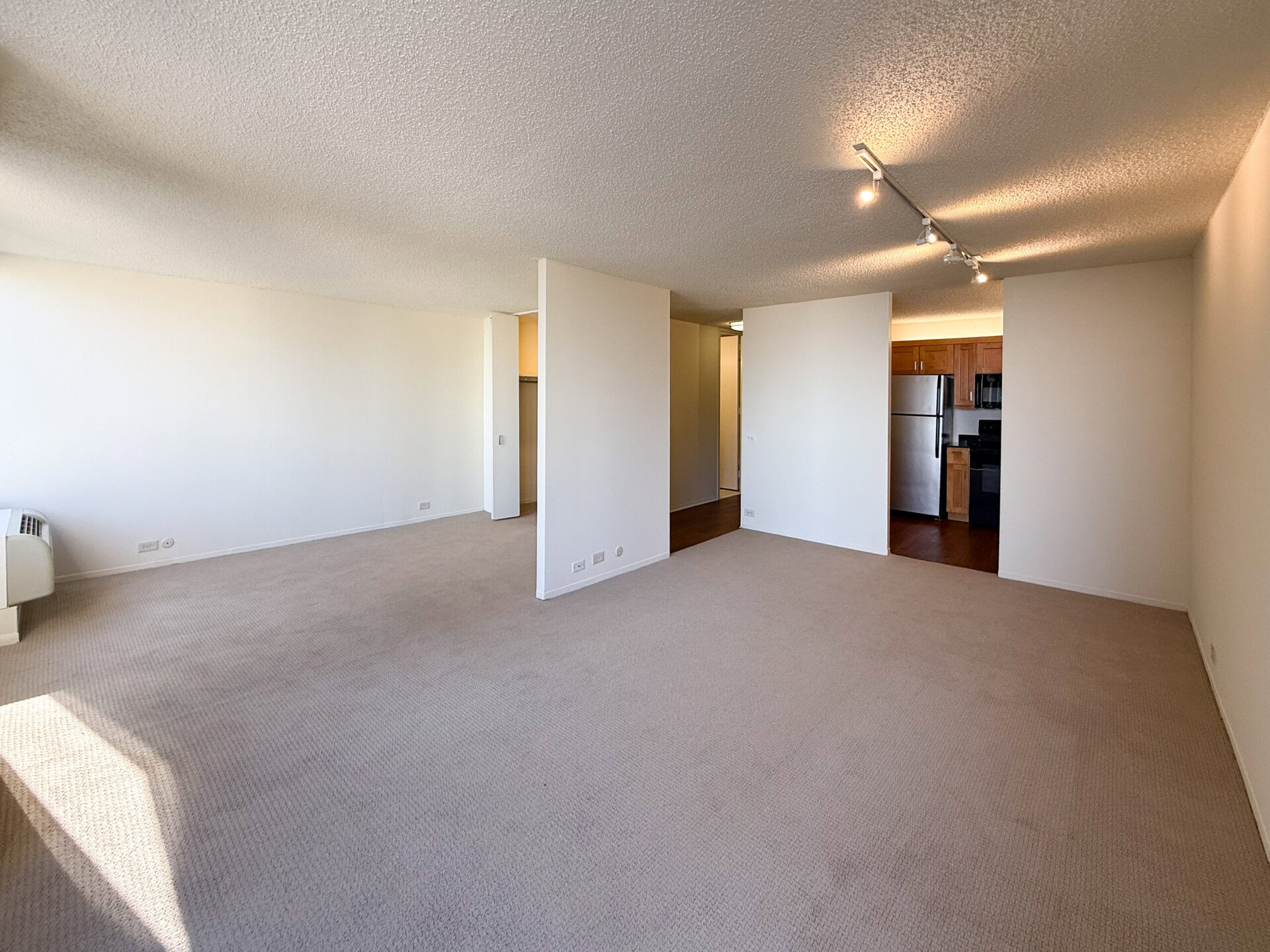 A spacious, empty living area with light beige carpet and textured ceiling. The walls are painted white, creating a bright atmosphere. On the left, there is an air conditioning unit, and to the right, a doorway leads to a kitchen area featuring wooden cabinets and stainless steel appliances, including a refrigerator. The layout suggests an open concept, with natural light illuminating the space.