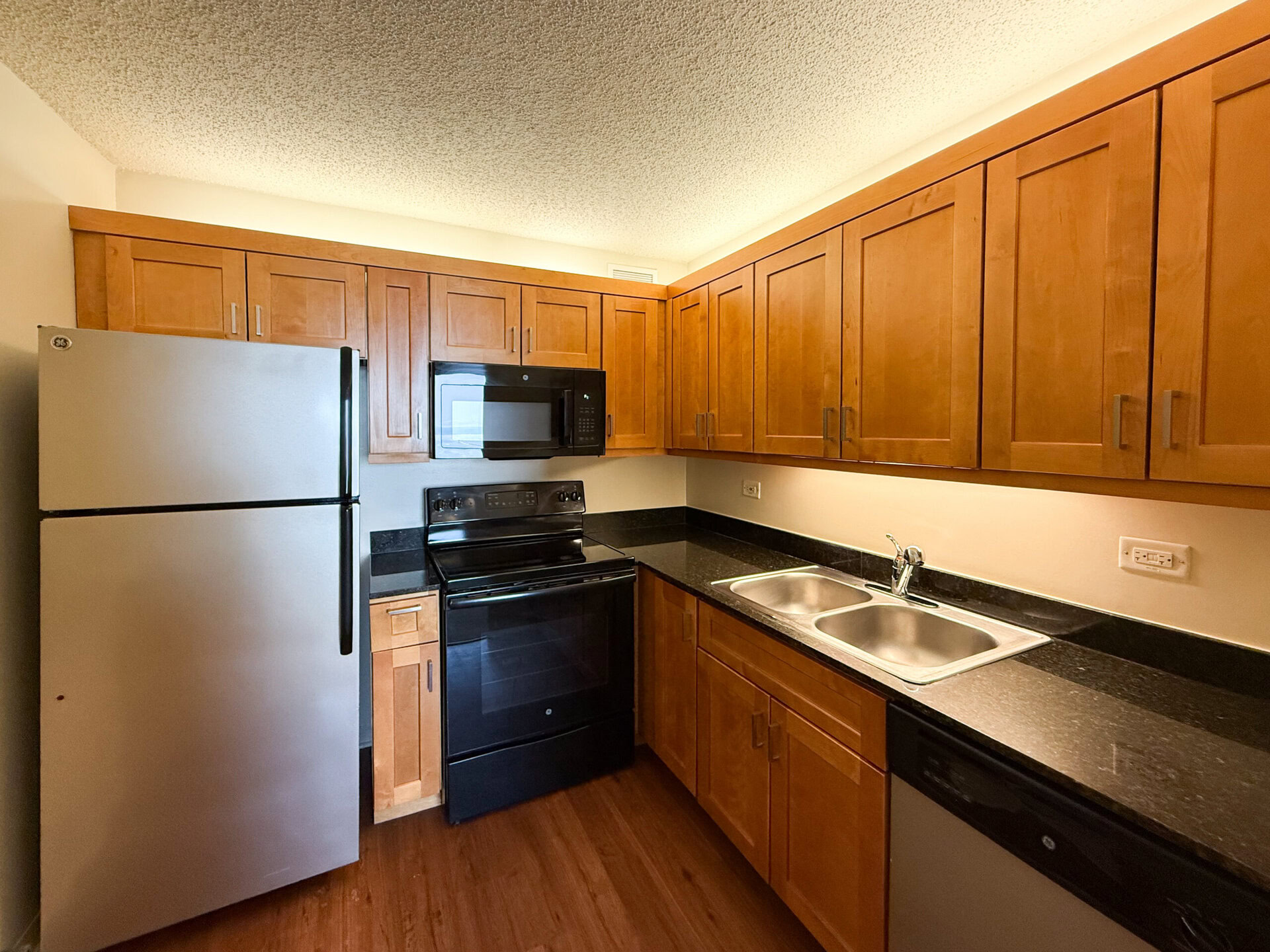 A modern kitchen featuring wooden cabinets with a warm finish, a black countertop, and stainless steel appliances. The appliances include a silver refrigerator, a black microwave mounted above a black electric stove, and a black dishwasher. A double sink is positioned next to the stove, with a sleek faucet. The overall design is functional and stylish, with ample storage space in the cabinets above.