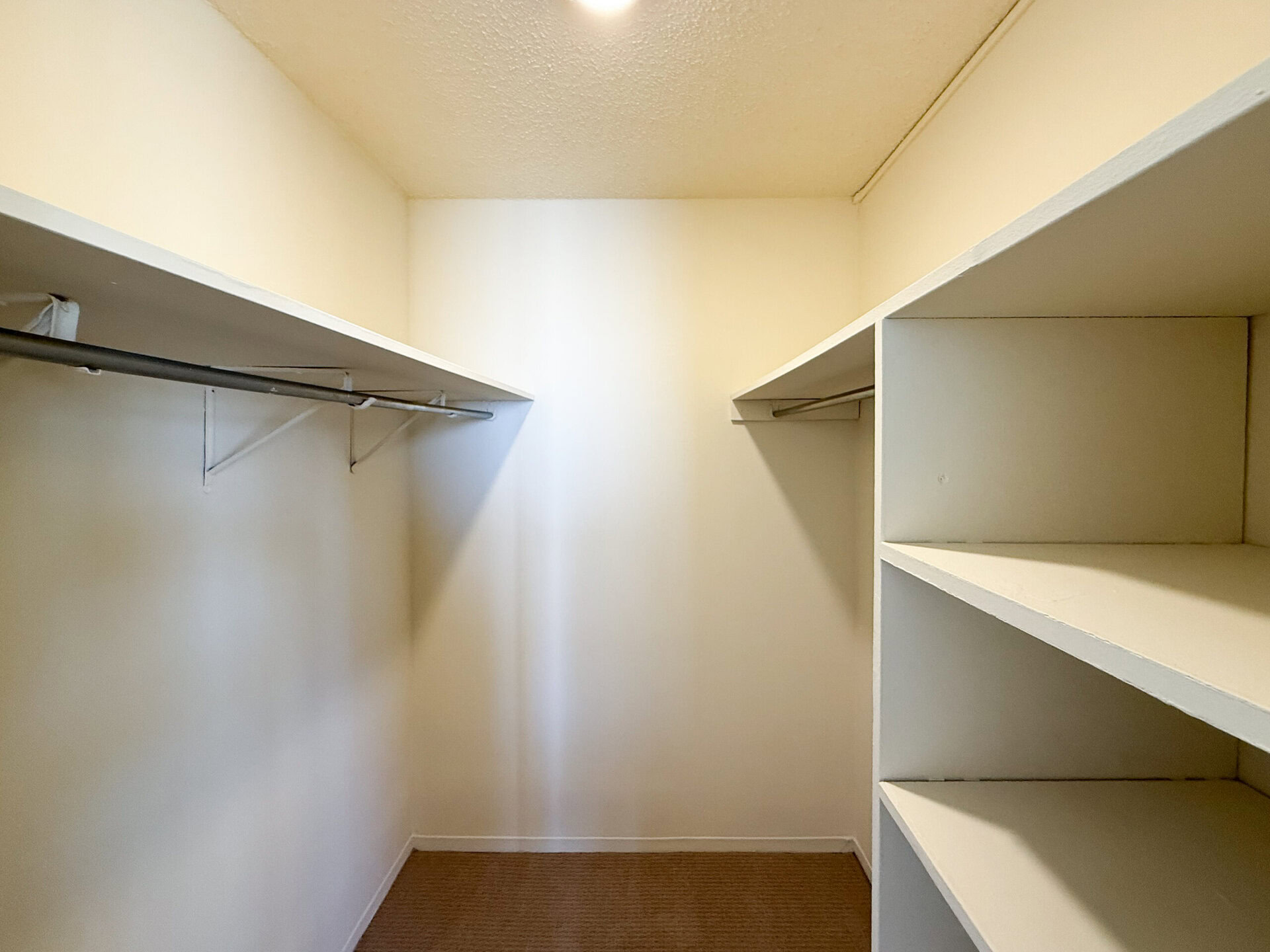 A spacious closet with light-colored walls features two horizontal shelves on either side. There are hanging rods supported by brackets along the back wall, providing ample space for clothing storage. The overall design is simple and functional, with a clean, uncluttered appearance.