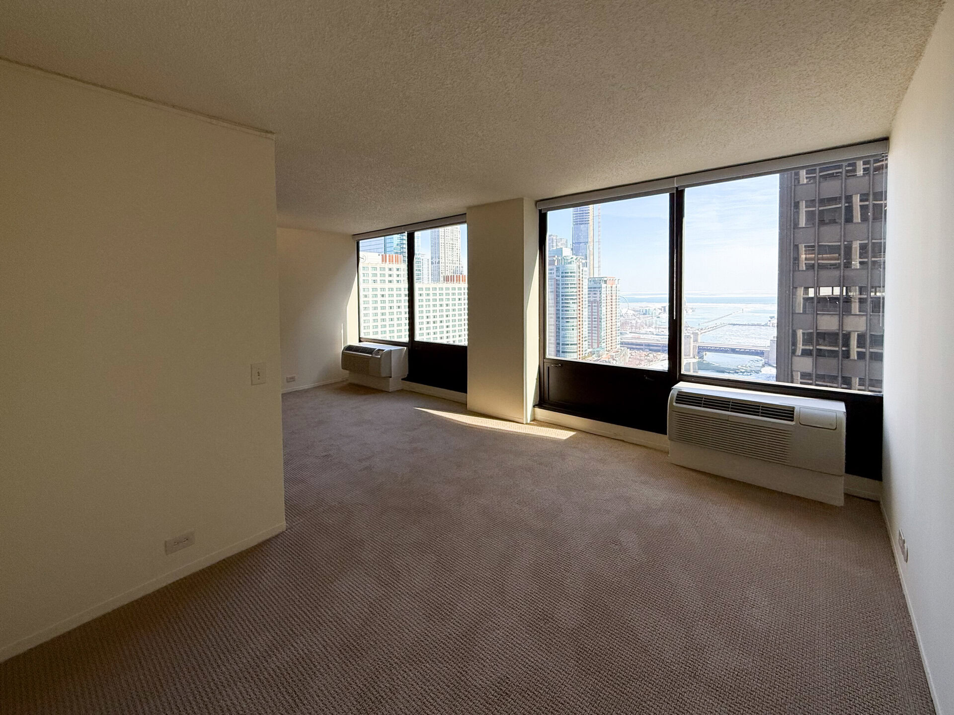A spacious, empty room with beige carpet and white walls. Large windows provide a view of a city skyline and a body of water in the distance. An air conditioning unit is installed beneath one of the windows. The room is well-lit, with natural light streaming in.