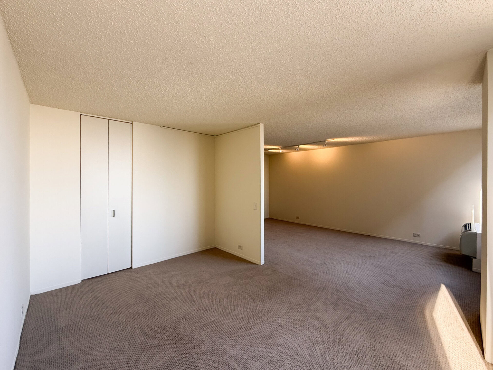 A spacious room with beige carpet and light-colored walls. On the left, there are double sliding doors leading to a closet. The room is divided by a partial wall, creating a sense of separation. The far side features a large, well-lit area with a window, and there is a small air conditioning unit visible on the right. The ceiling has a textured finish, and the overall ambiance is bright and open.