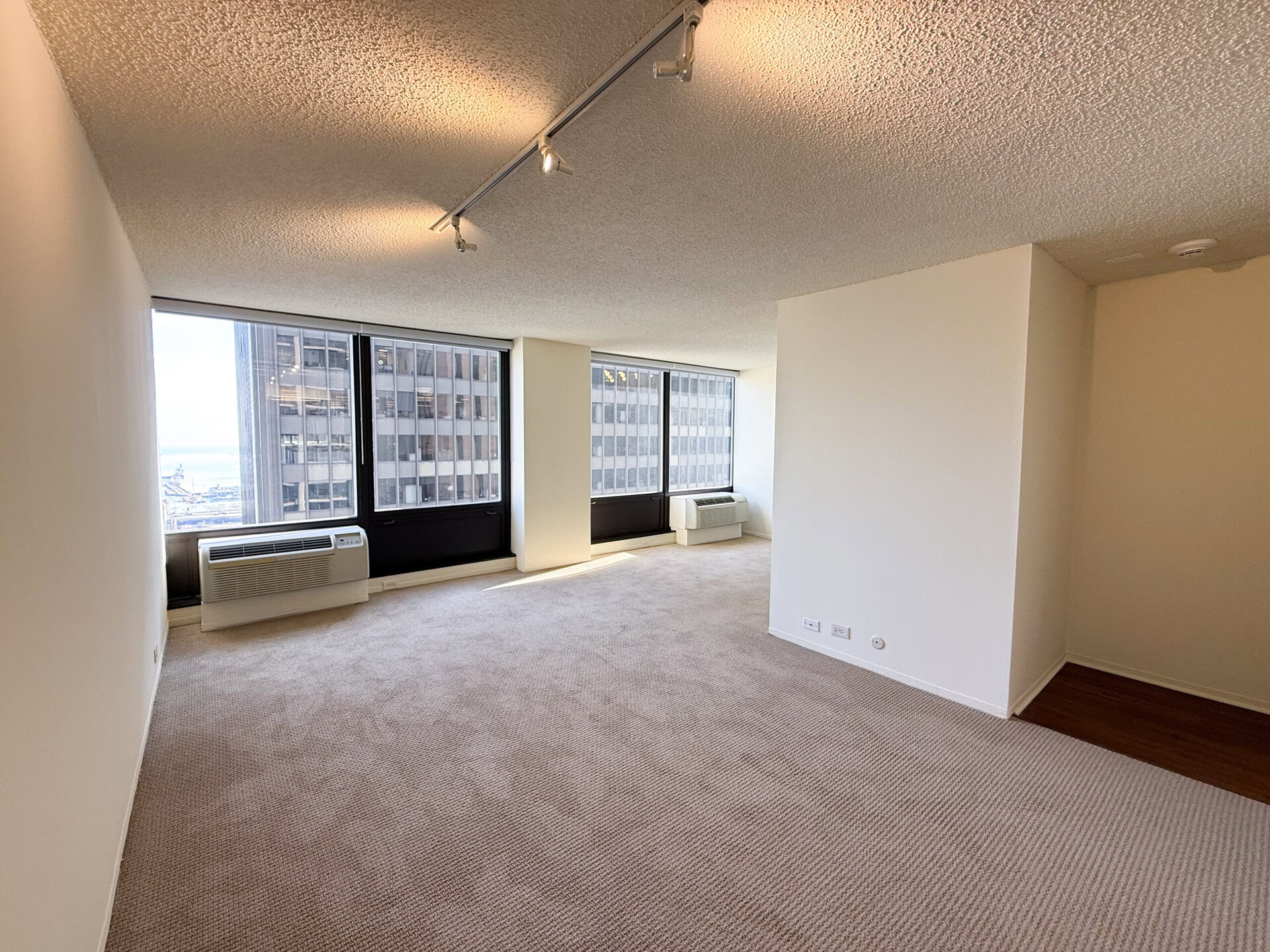 A spacious, empty room with beige carpet and white walls. Large windows provide a view of nearby buildings and the horizon. Two air conditioning units are positioned beneath the windows. The ceiling features track lighting, and there is a corner with hardwood flooring visible. The overall atmosphere is bright and airy, with natural light flooding the space.