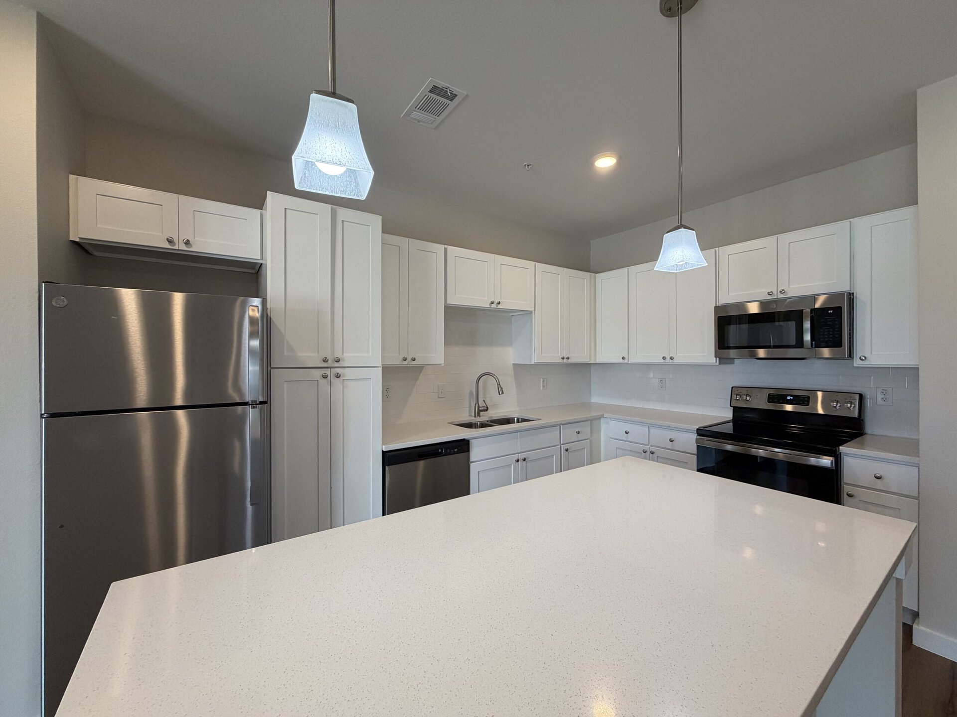 A modern kitchen featuring white cabinetry and stainless steel appliances. The kitchen includes a large island with a white countertop, a stainless steel refrigerator, a black oven, and a microwave above it. A double sink is positioned under a window, with a sleek faucet. Two pendant lights hang above the island, providing illumination. The backsplash is white tile, complementing the overall clean and contemporary design.