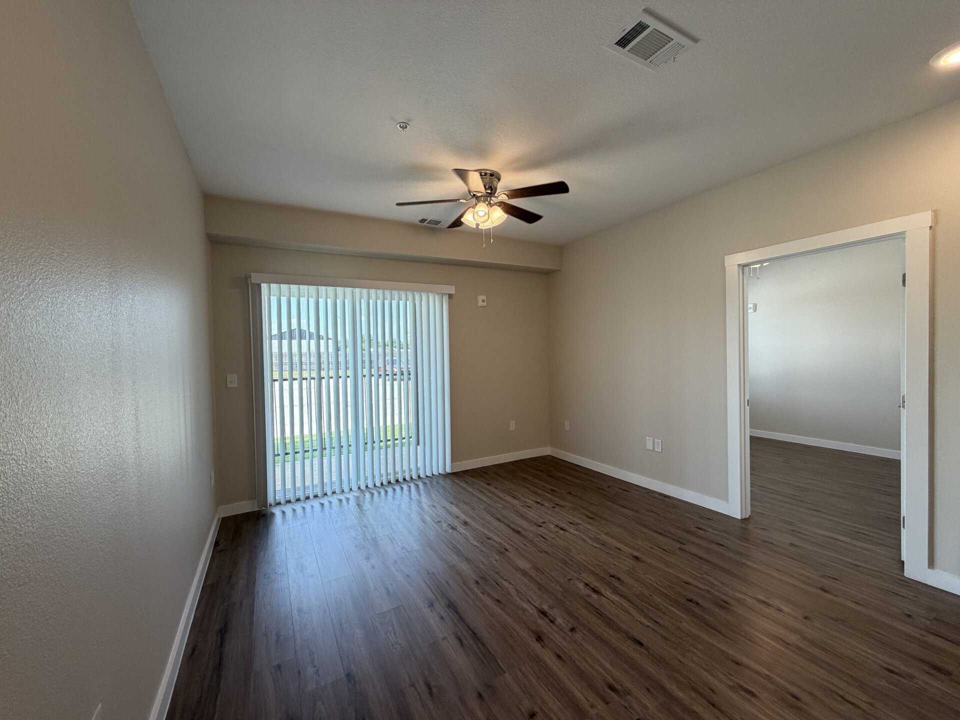 A spacious room features light-colored walls and a ceiling fan with three lights. The floor is made of wood-like laminate. On one side, there are vertical blinds covering a sliding glass door that leads outside, allowing natural light to enter. To the right, an open doorway leads to another room, enhancing the sense of space. The overall ambiance is bright and inviting.