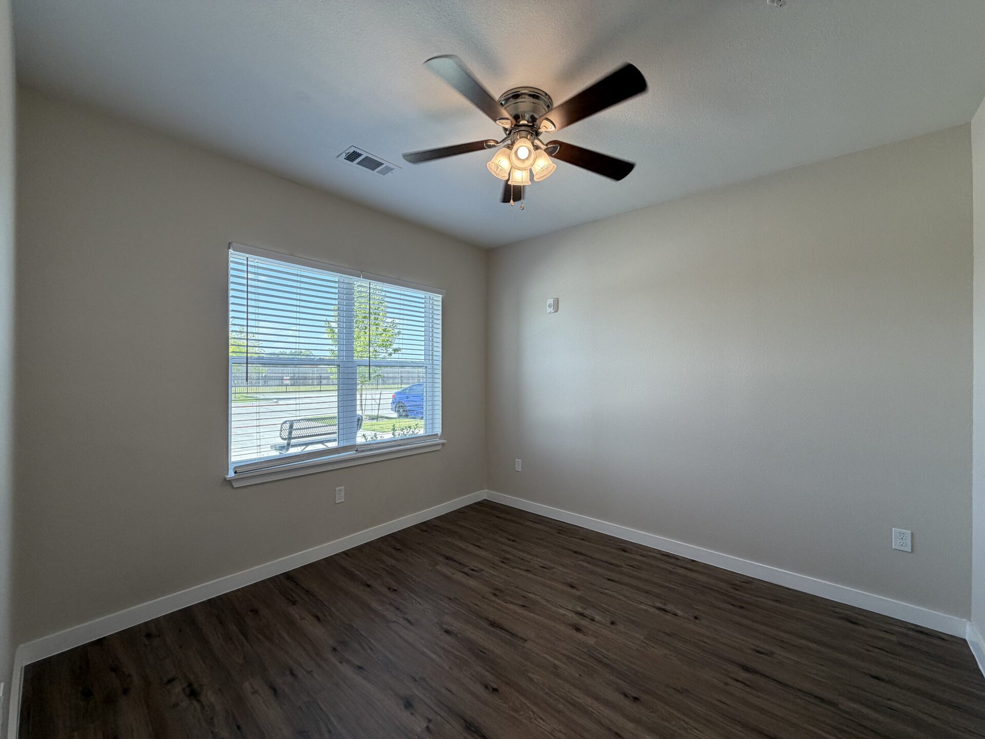 A well-lit room features a ceiling fan with multiple light fixtures. The walls are painted a soft beige, and the floor has a wood-like finish. A large window with horizontal blinds allows natural light to enter, revealing a view of a tree and a bench outside. The room is empty, providing a spacious feel.