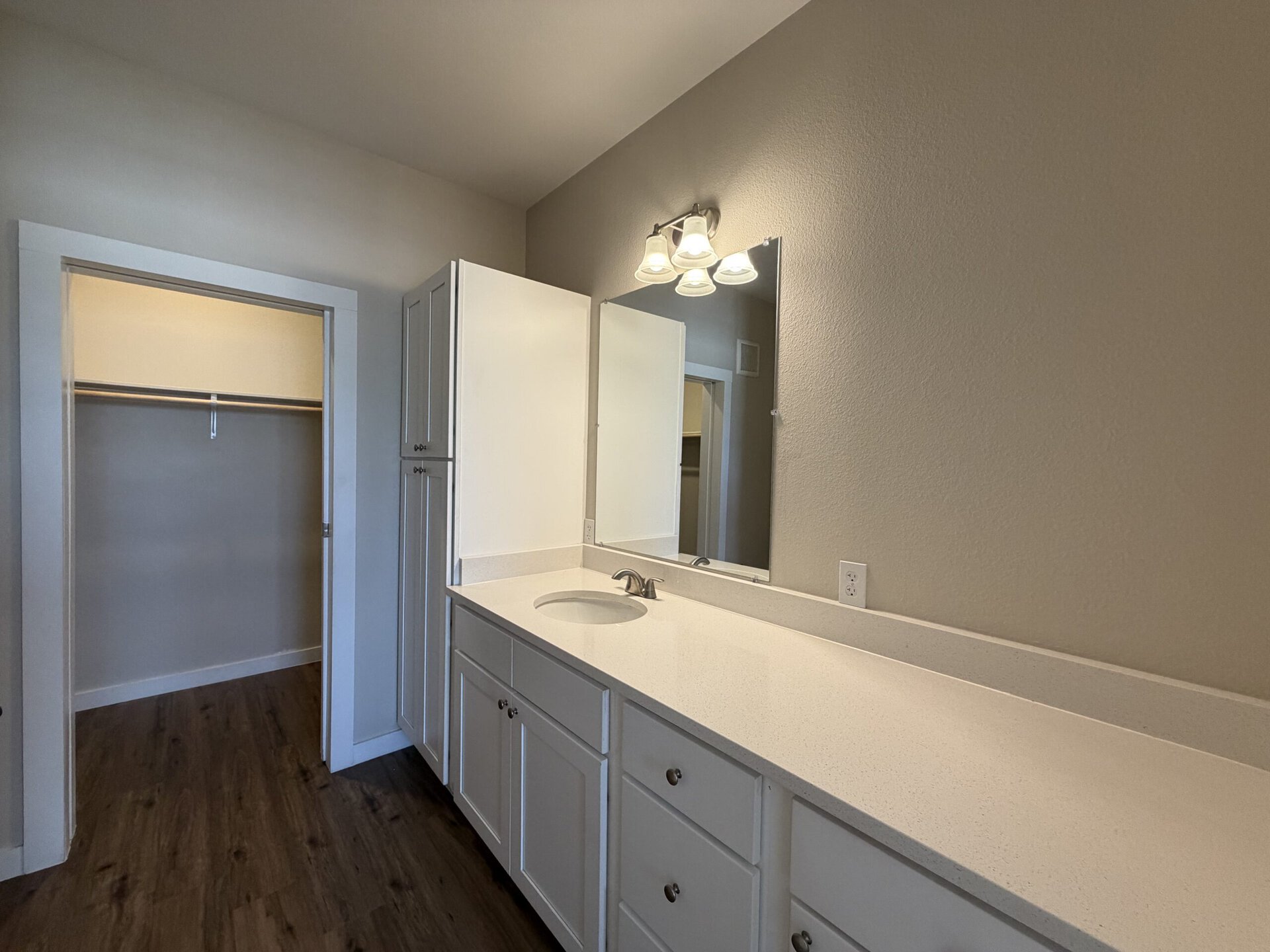 A modern bathroom features a white countertop with a circular sink and a sleek faucet. Above the sink, there is a large mirror and three light fixtures providing illumination. To the left, a tall cabinet with multiple drawers and doors offers storage space. An open closet is visible in the background, showcasing a simple design with a hanging rod. The walls are painted in a soft neutral color, and the flooring is a dark wood finish, creating a warm and inviting atmosphere.