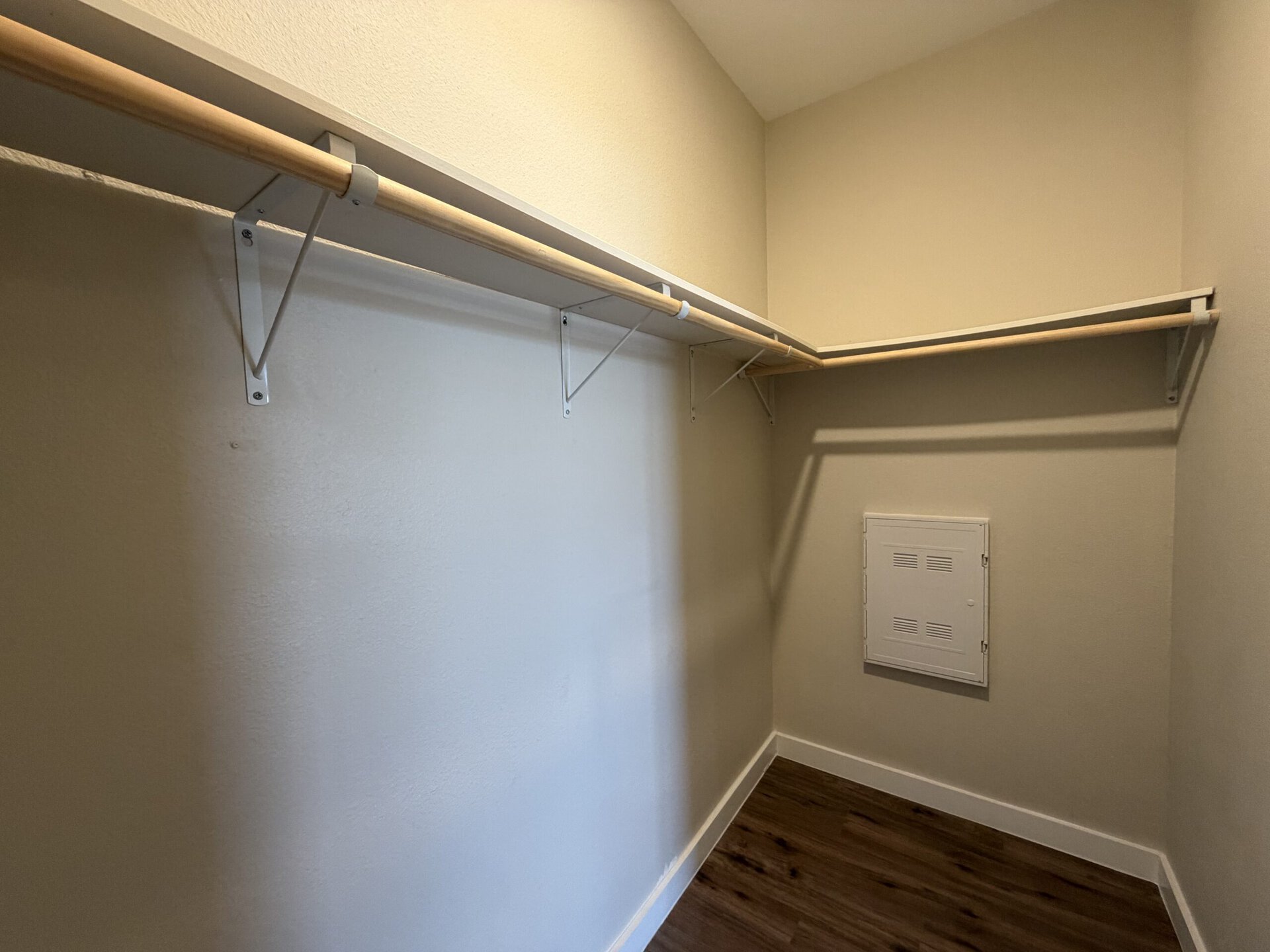 A small closet features a wooden hanging rod supported by white brackets on one side. The opposite wall is bare, with a small white vent cover mounted at the lower right. The walls are painted a light color, and the floor appears to be a dark wood laminate. The overall space is uncluttered and well-lit.