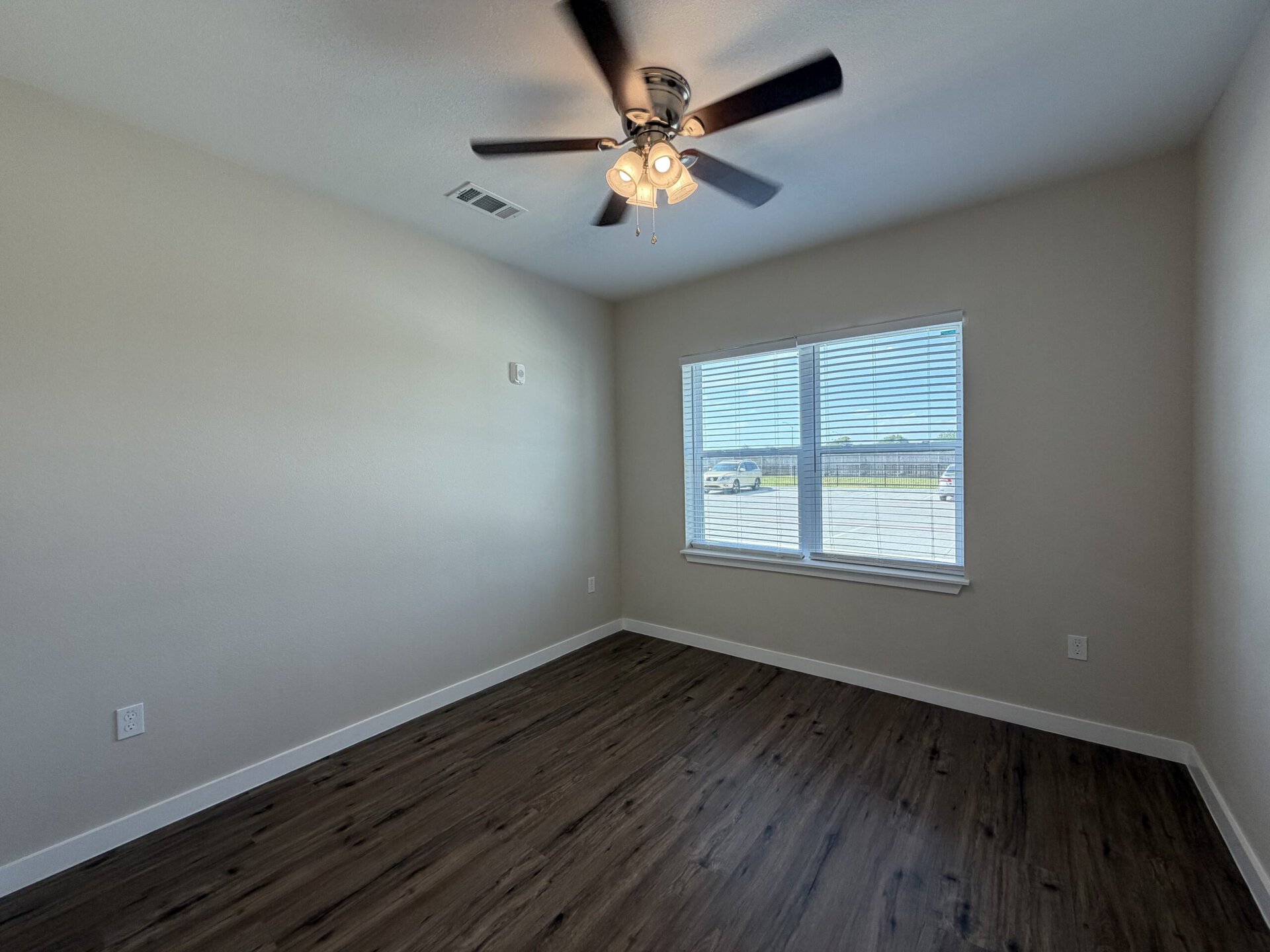A small, well-lit room features light-colored walls and a dark wood floor. A ceiling fan with three light fixtures hangs from the ceiling. On one side, there is a large window with horizontal blinds, allowing natural light to enter and providing a view of an outdoor area. The room appears empty and spacious, with a power outlet visible on one wall.