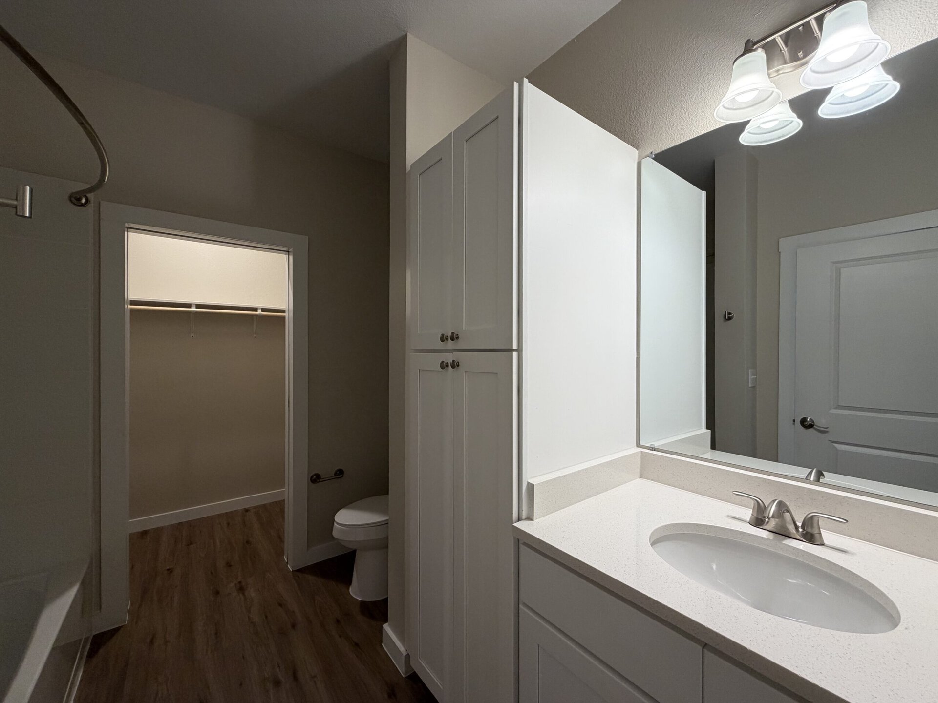 A modern bathroom features a white vanity with a sink and a sleek faucet. Above the sink, there is a large mirror and three light fixtures providing illumination. To the left, a doorway leads to a closet with a hanging rod. A toilet is positioned nearby, and the flooring is a wood-like laminate. The walls are painted in a neutral tone, enhancing the bright and clean aesthetic of the space.