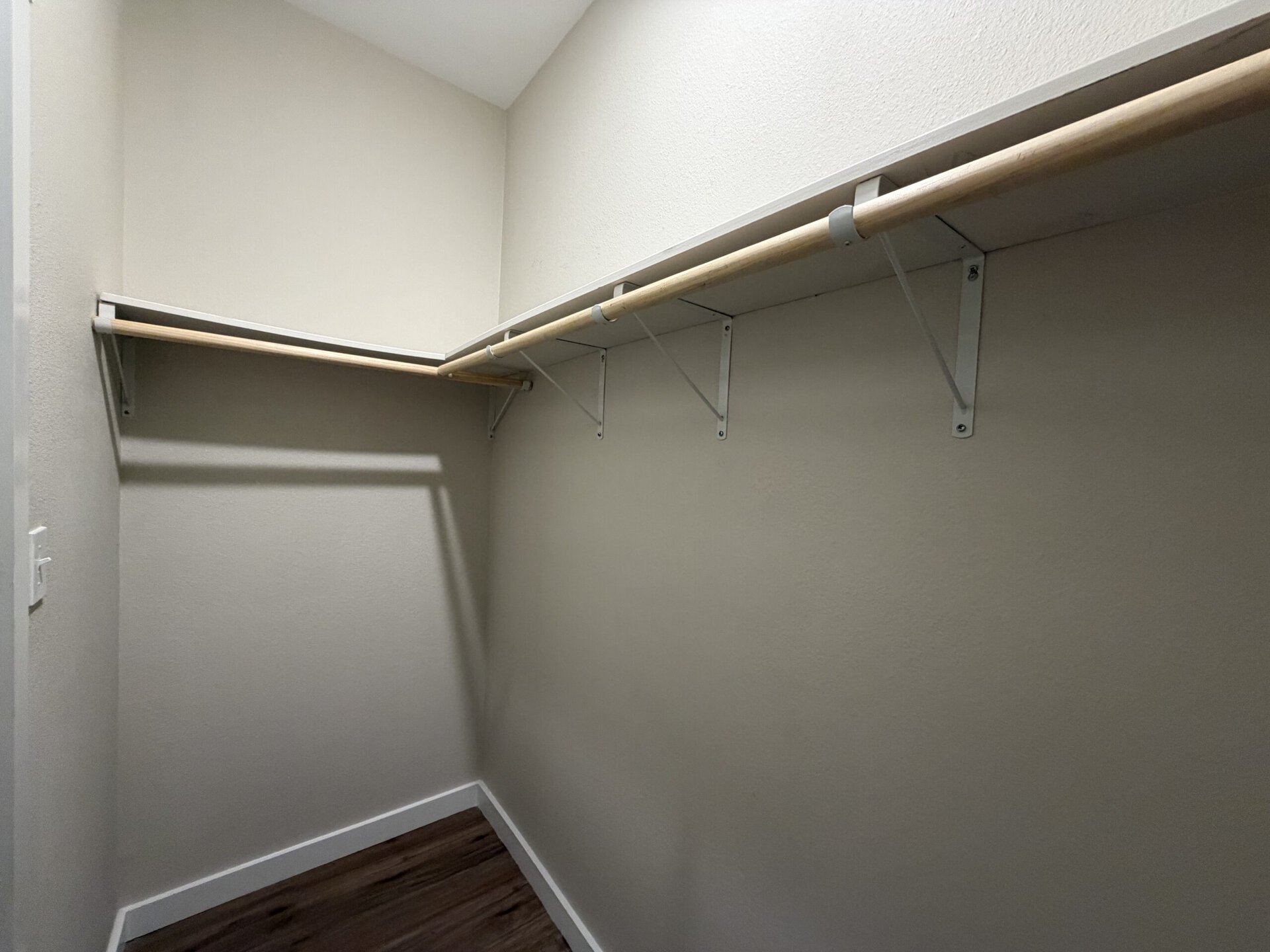 A small, empty closet features a single wooden hanging rod supported by white metal brackets. The walls are painted a light color, and the floor appears to have a wood-like finish. There is a shelf above the rod, also supported by the same brackets, providing additional storage space. The overall space is minimalistic and uncluttered.