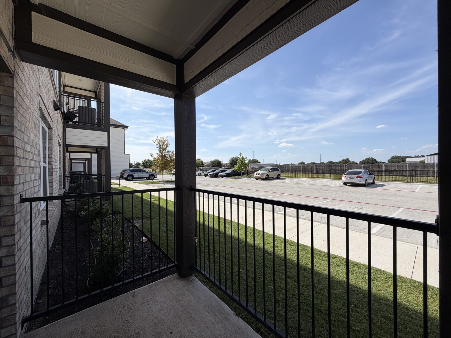 A view from a balcony shows a well-maintained apartment complex with brick exteriors. The foreground features a black railing and a small patch of grass with some shrubs. In the background, a parking lot is visible with several parked cars, including a silver sedan. The sky is clear with a few clouds, and there are trees lining the area, contributing to a pleasant outdoor atmosphere.