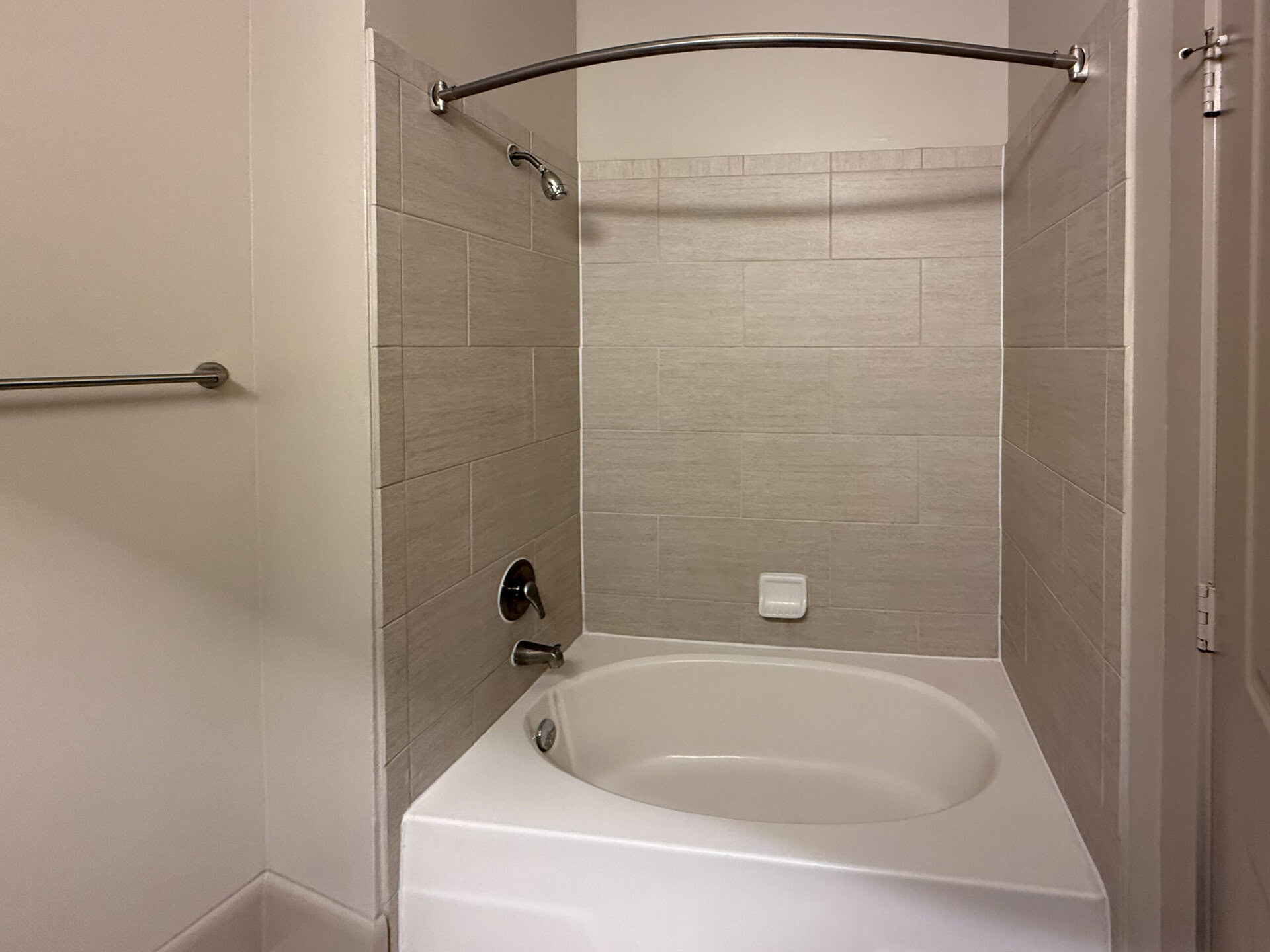 A modern bathroom features a white, oval bathtub set against light gray, textured wall tiles. The tub has a chrome faucet and a showerhead mounted on the wall above. A grab bar is installed on the adjacent wall, and a small soap dish is positioned on the wall near the tub. The overall design is clean and minimalistic, with neutral colors creating a calming atmosphere.