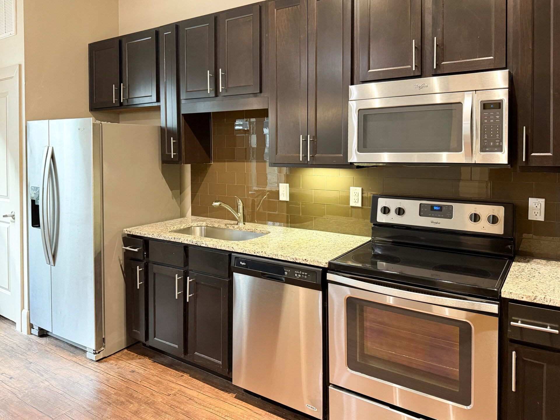A modern kitchen featuring dark wood cabinetry and a light granite countertop. On the left, there is a stainless steel refrigerator with a water dispenser. The sink, positioned centrally, has a sleek faucet. Below the countertop, a stainless steel dishwasher is visible. To the right, an electric stove with an oven is installed, accompanied by a stainless steel microwave above it. The backsplash is made of glossy brown tiles, and the kitchen is well-lit with warm lighting.